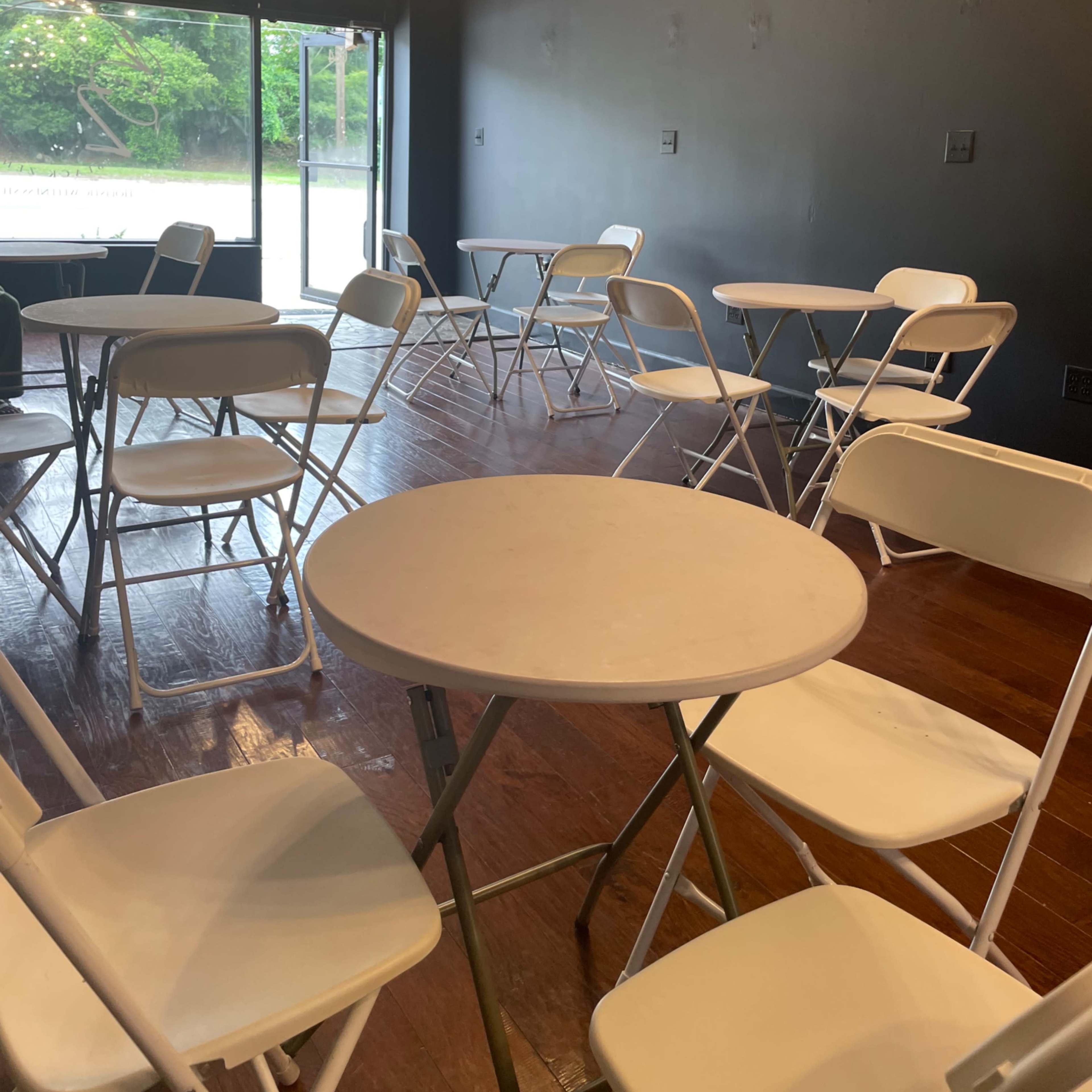 A room with several white folding tables and chairs arranged on a hardwood floor, featuring a large window overlooking an outdoor area.
