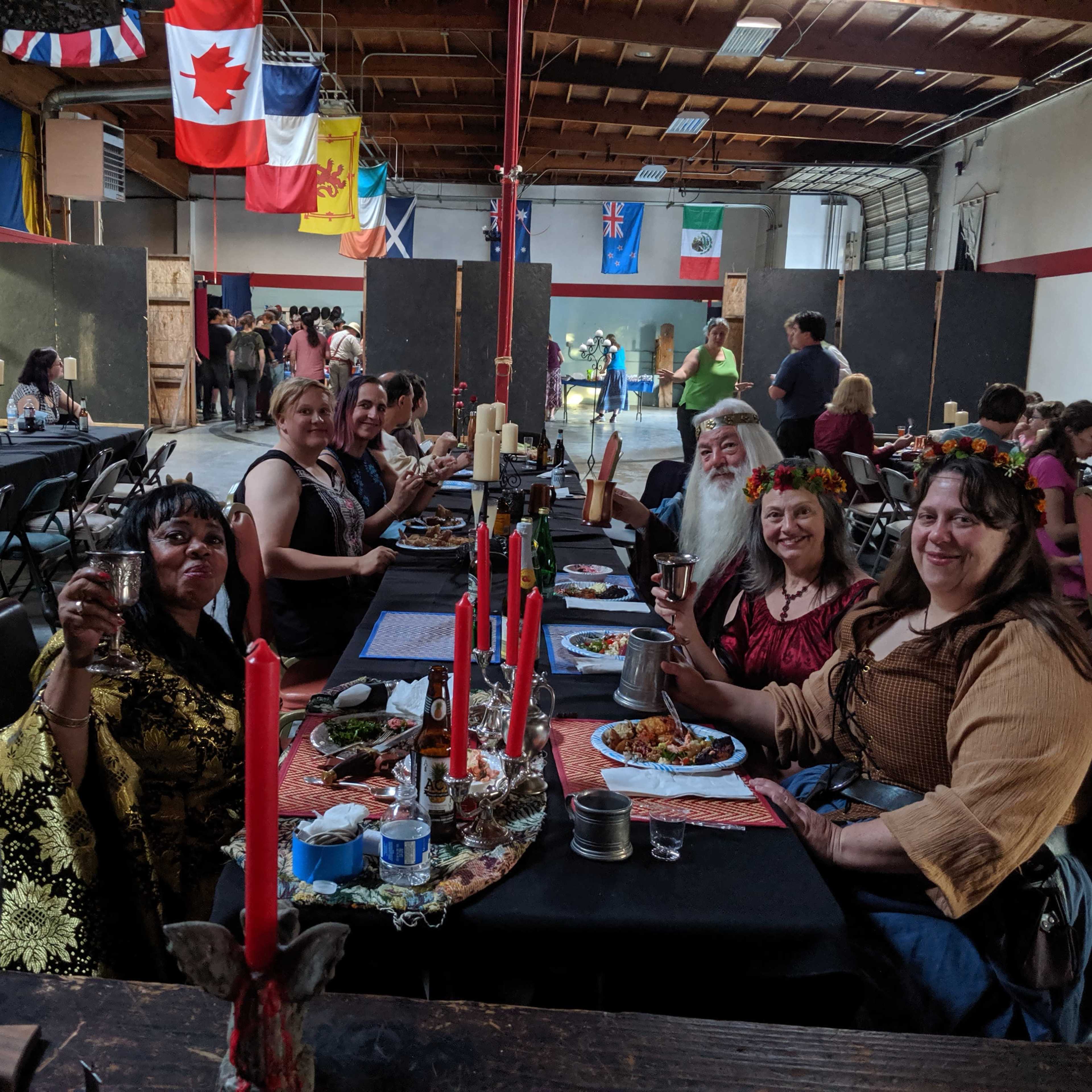 A group of people sits at a long table adorned with candles and food, while flags hang from the ceiling in a spacious hall.