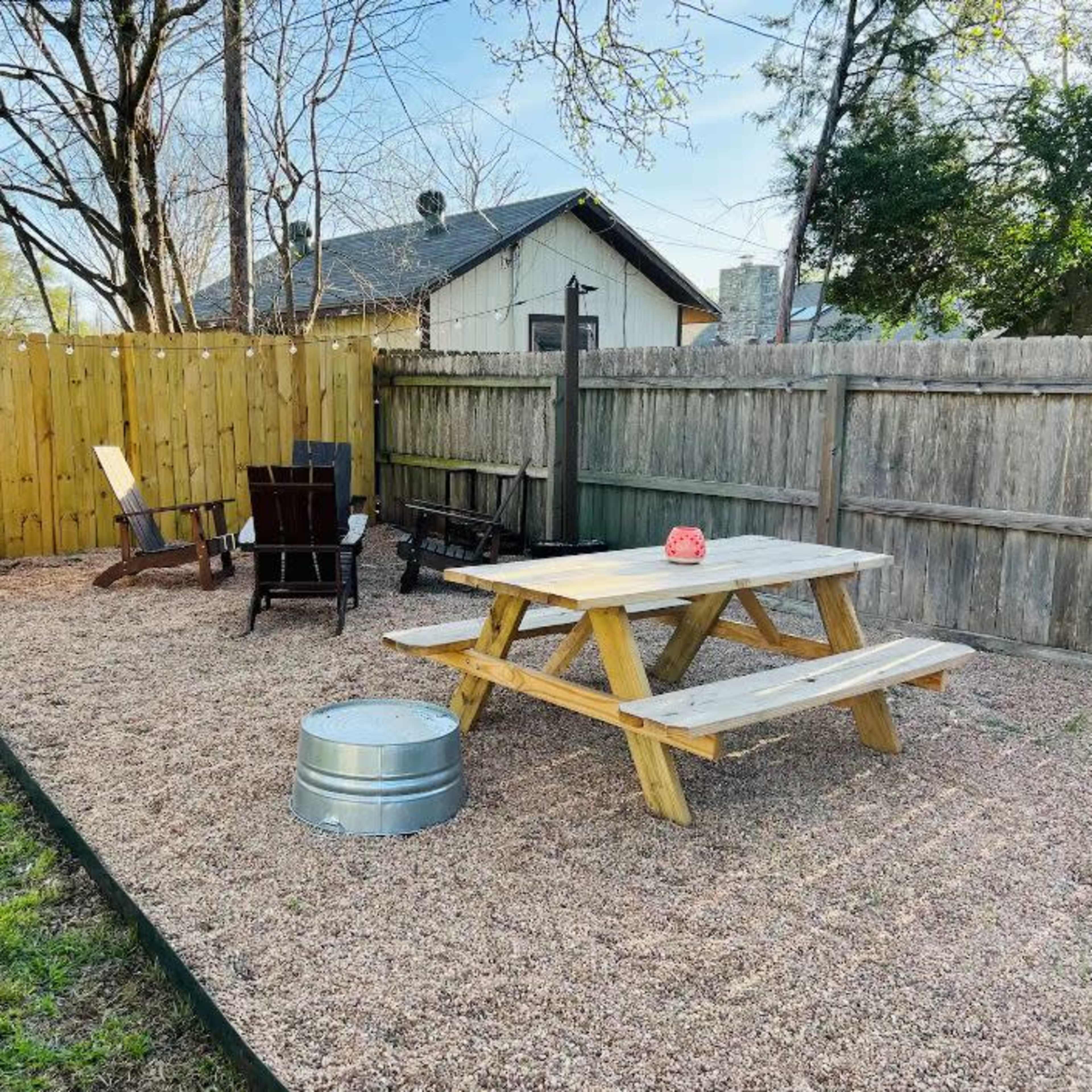 A gravel-covered outdoor space with a wooden picnic table, two black chairs, and a metal fire pit surrounded by a wooden fence and trees.