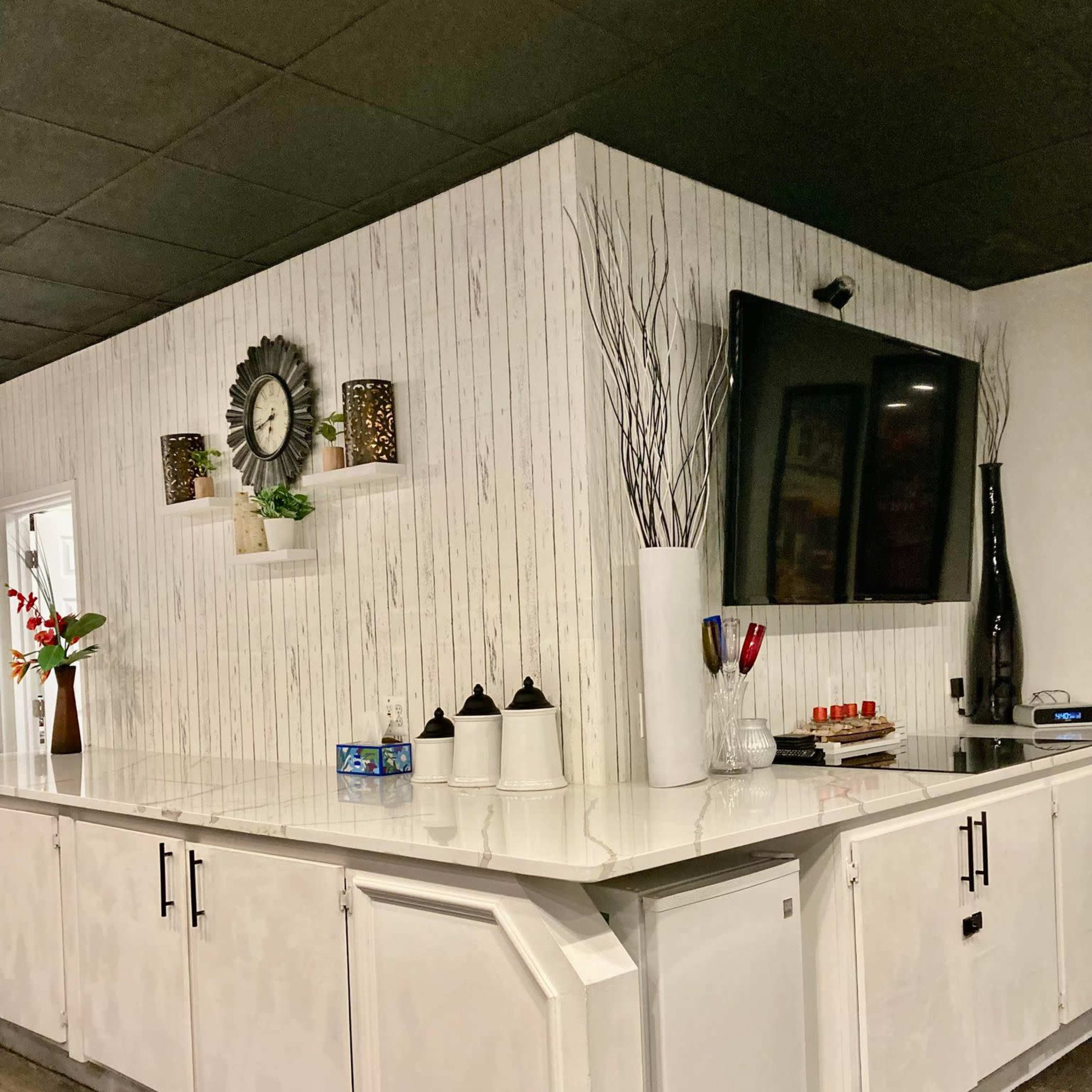 A modern kitchen area with a white countertop, a large flat-screen TV mounted on the wall, and decorative items on shelves.