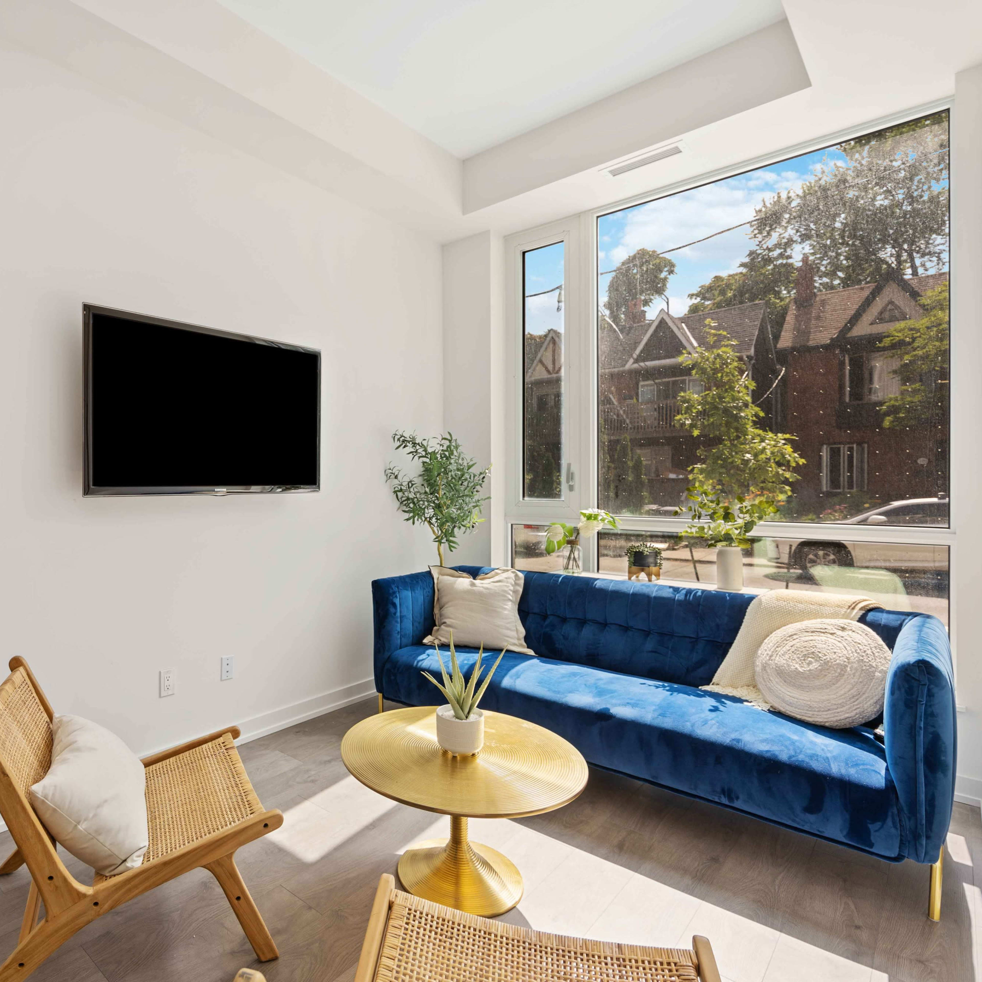 A small living room features a blue couch, a round gold table, and two chairs, with a television mounted on the wall and large windows allowing natural light.