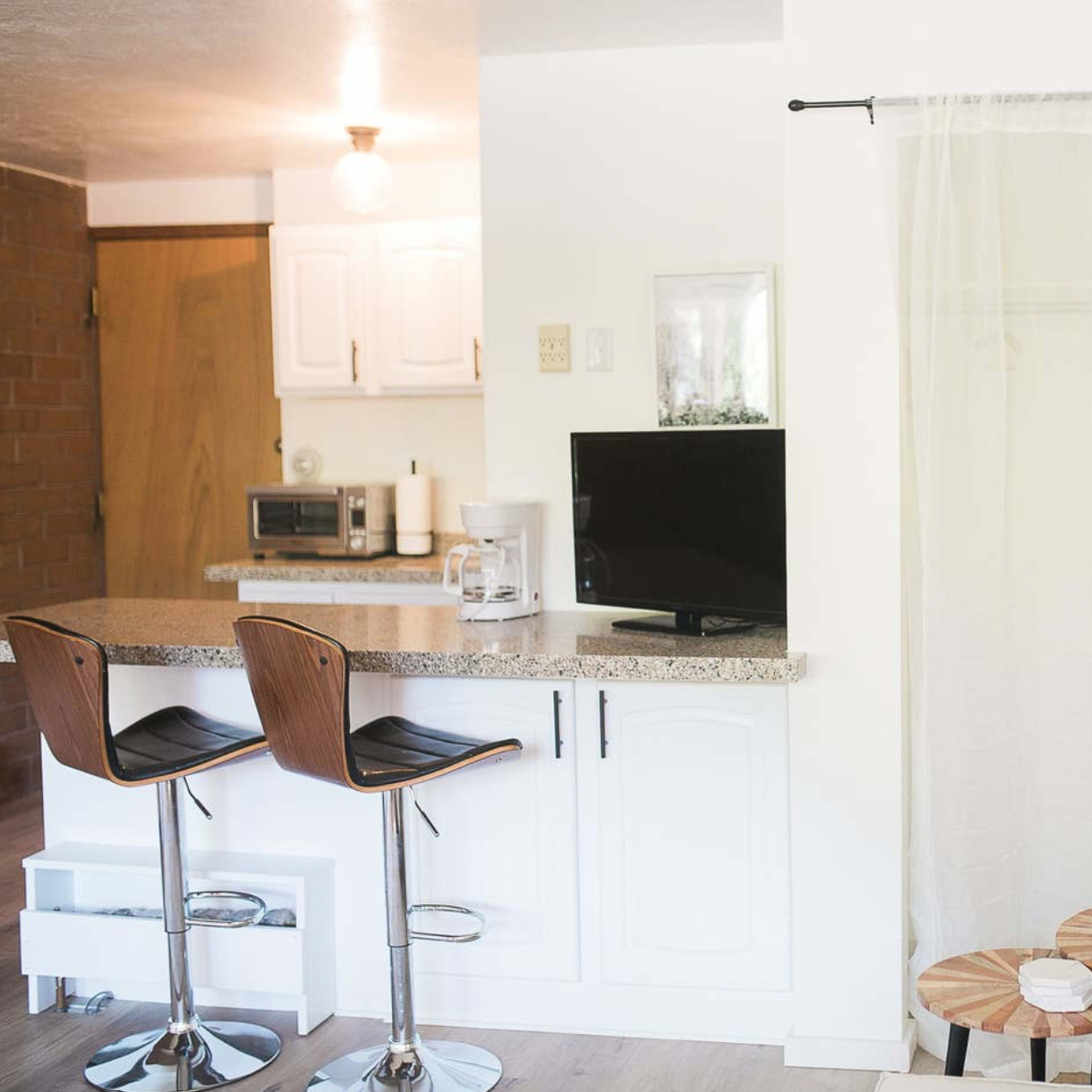 A bright, modern living space with a kitchenette featuring a bar counter and two stools, a small TV on the counter, and a curtain separating the area from a seating space.