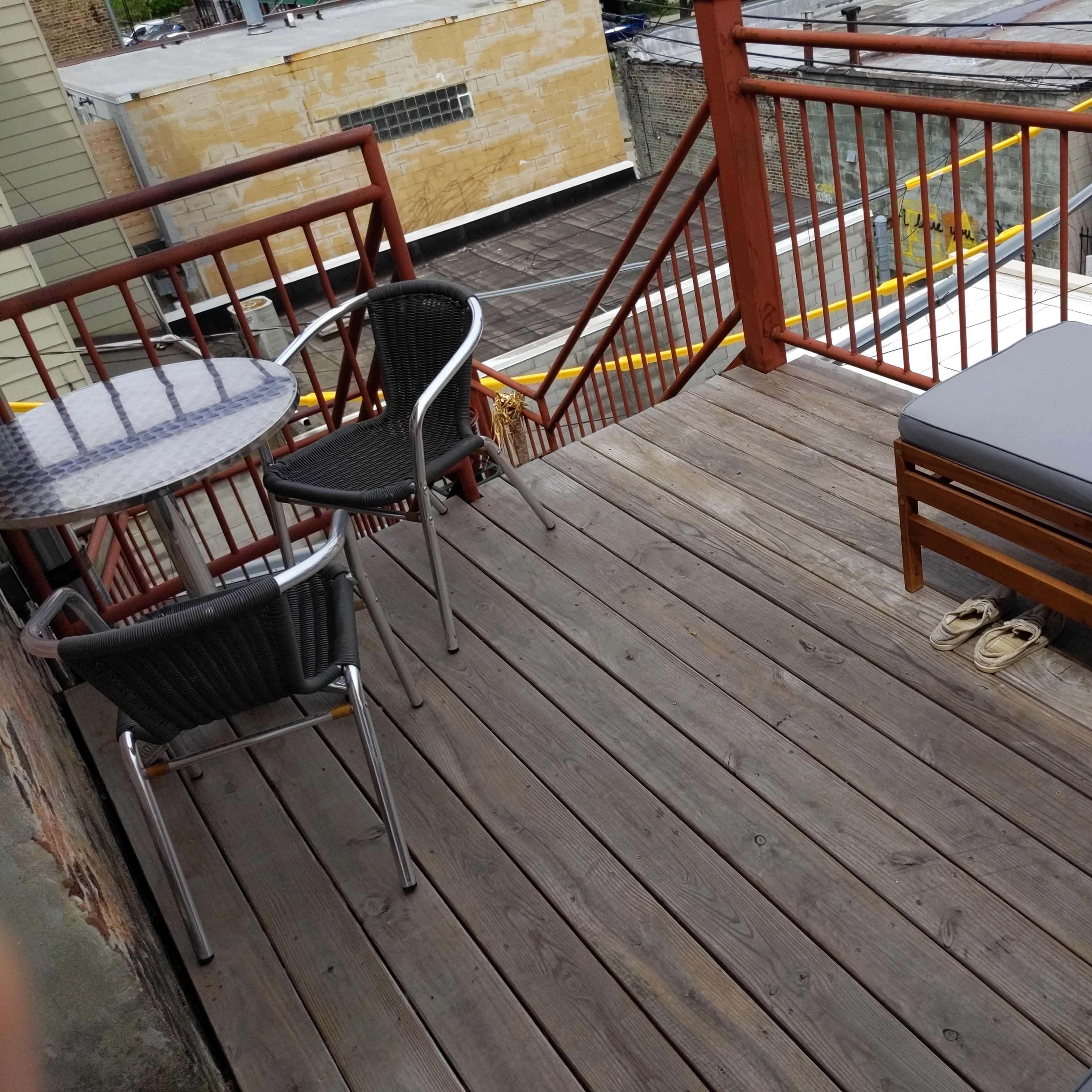 A wooden deck features a round table with two chairs and a sectional couch, with a pair of sandals resting on the floor.