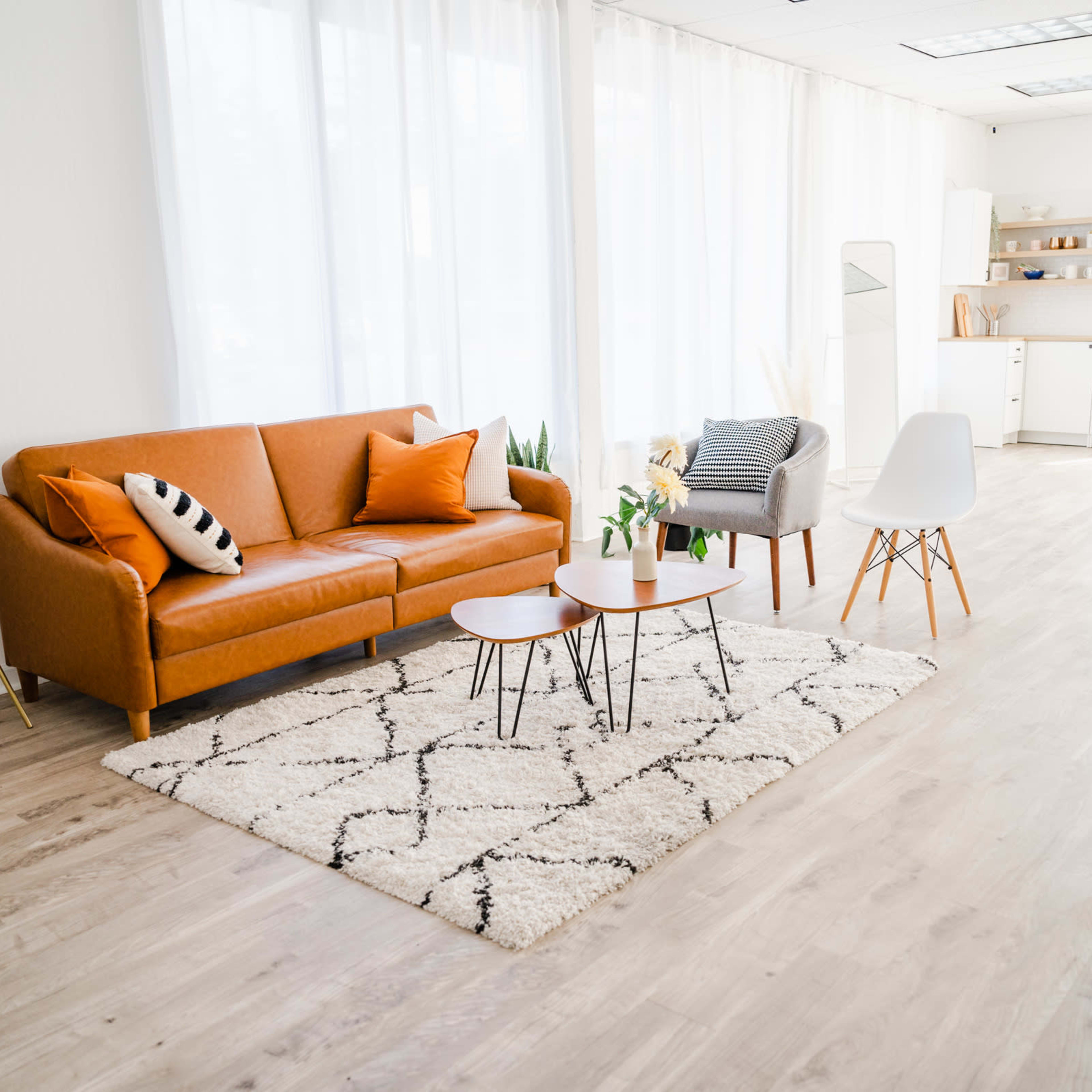A bright, modern living space featuring an orange sofa, two coffee tables, and a mix of decorative chairs, with large windows allowing natural light to fill the room.