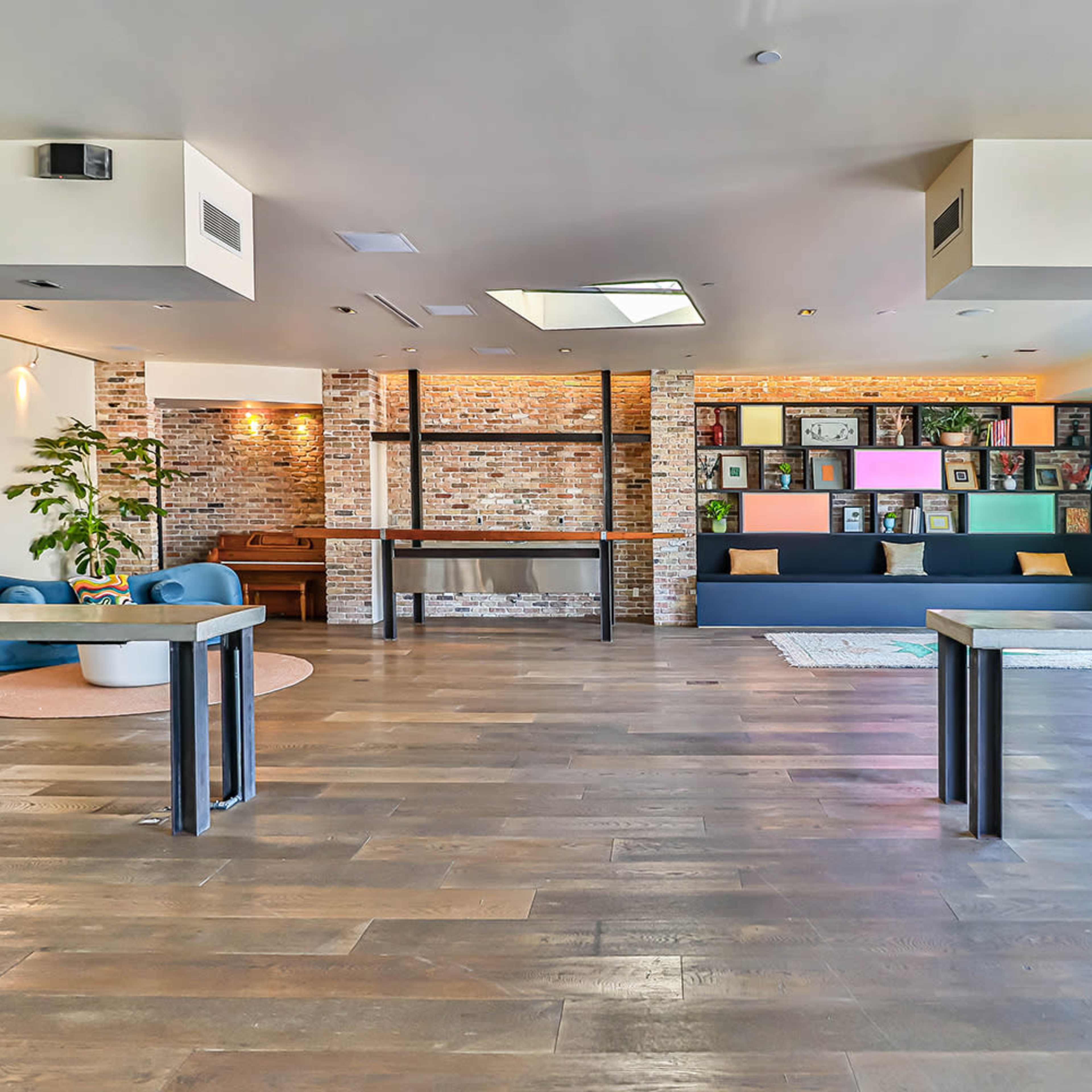 A spacious interior featuring wooden floors, blue seating, a brick wall, and large windows allowing natural light.
