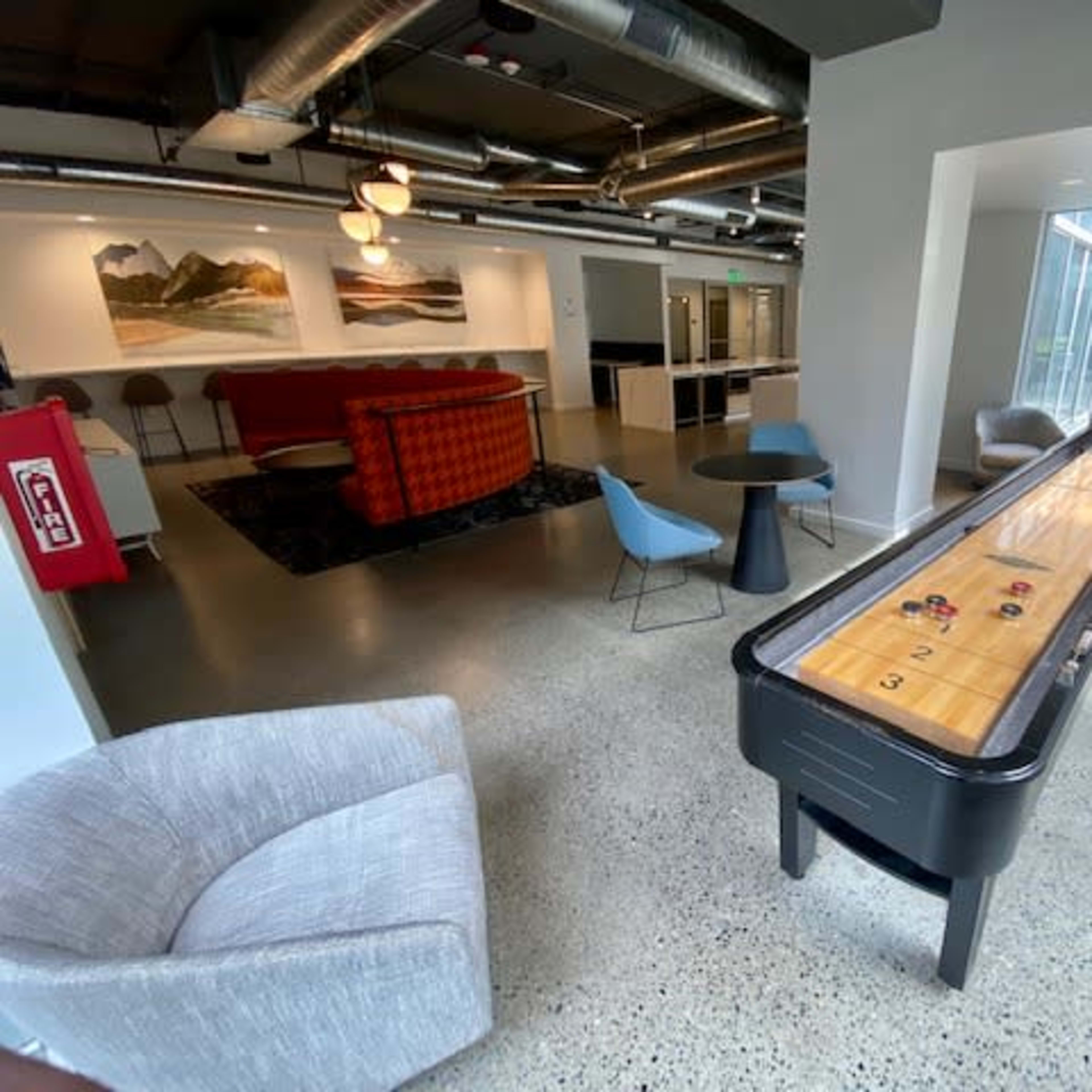 A modern lounge area with a shuffleboard table, comfortable seating, and large windows letting in natural light.