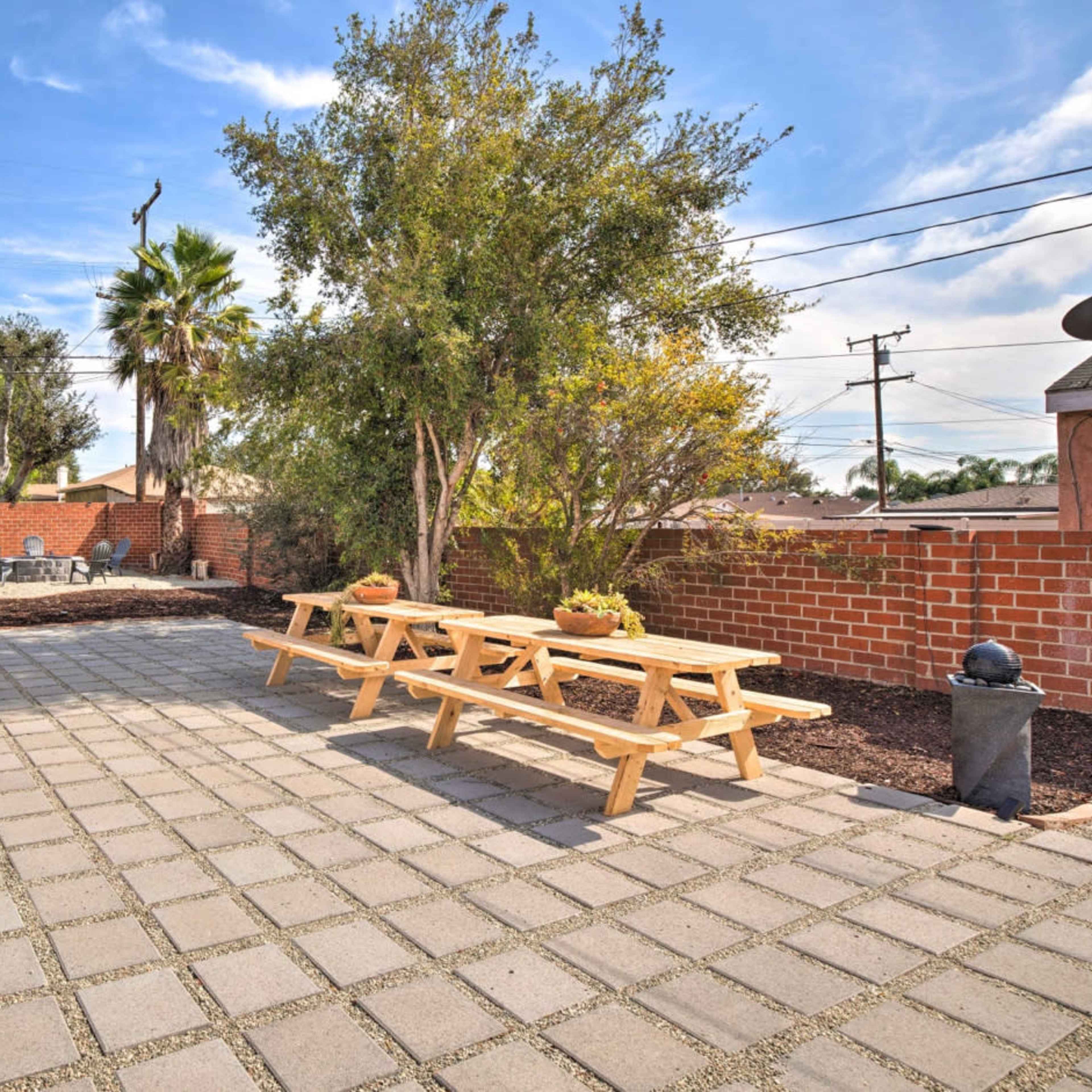 A spacious outdoor patio area with stone pavers, a wooden picnic table set, and landscaped greenery surrounded by a brick wall.
