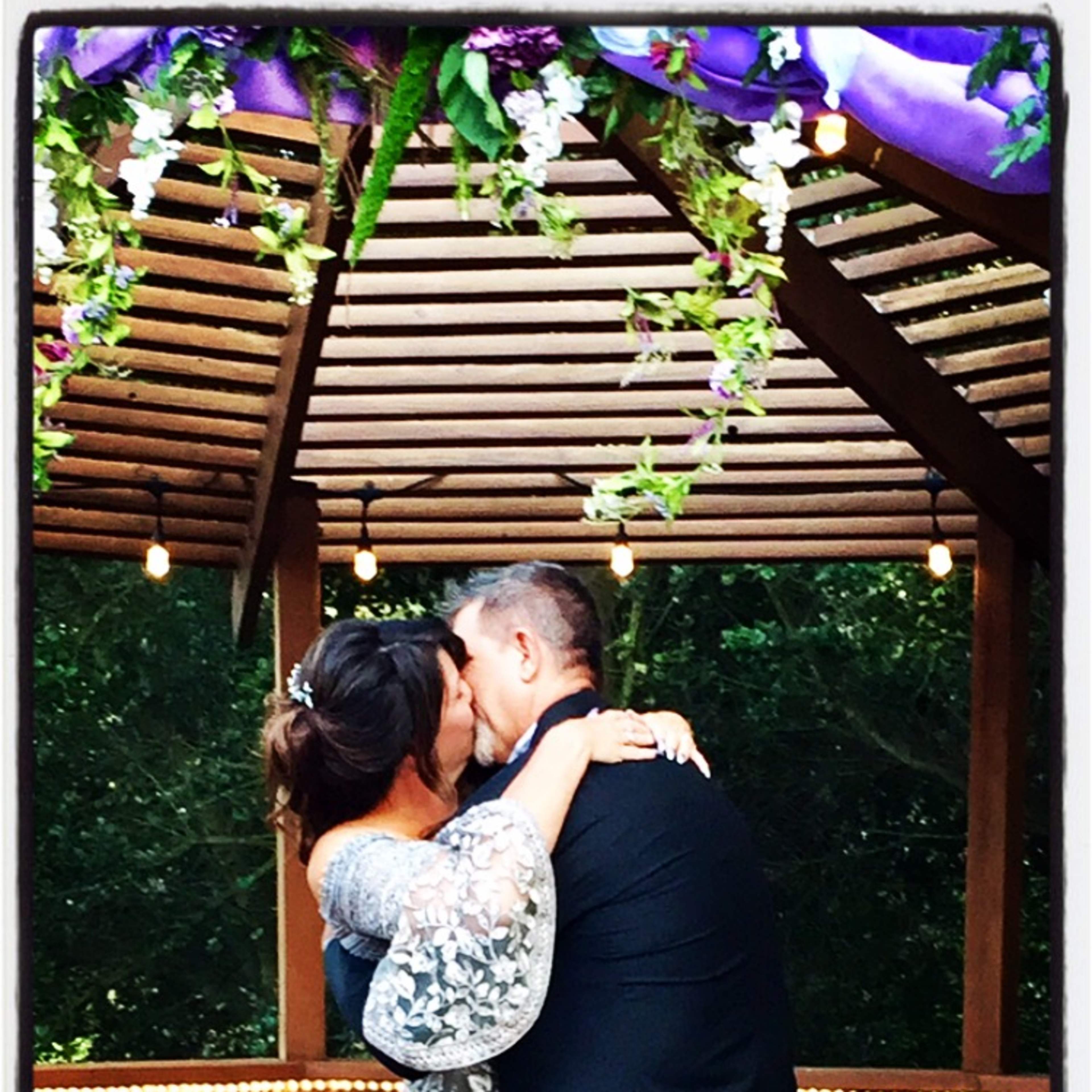 A couple kisses under a decorated gazebo with hanging flowers and string lights.