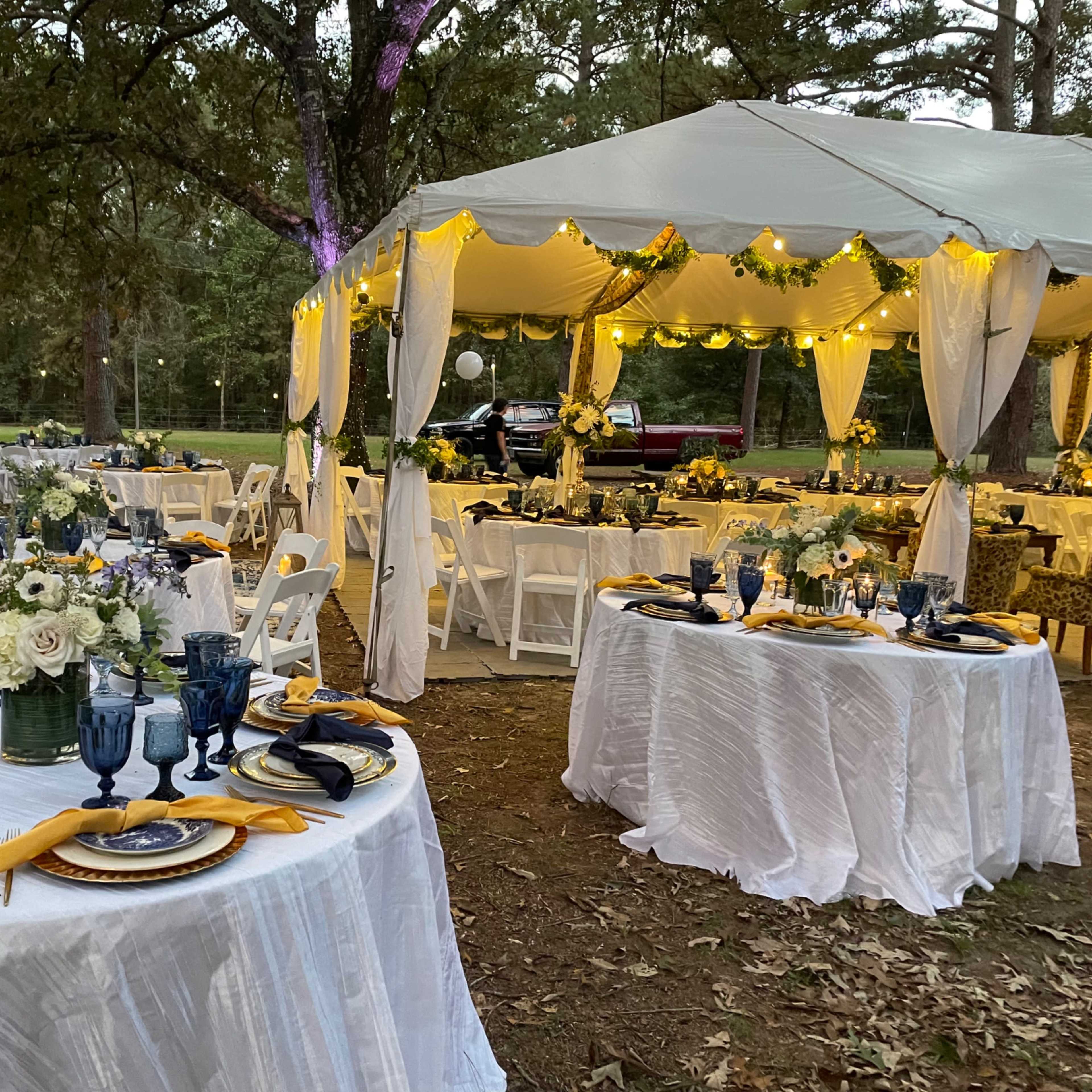 A decorative outdoor dining setup with white tablecloths, blue glassware, floral centerpieces, and a large covered tent set amidst trees.