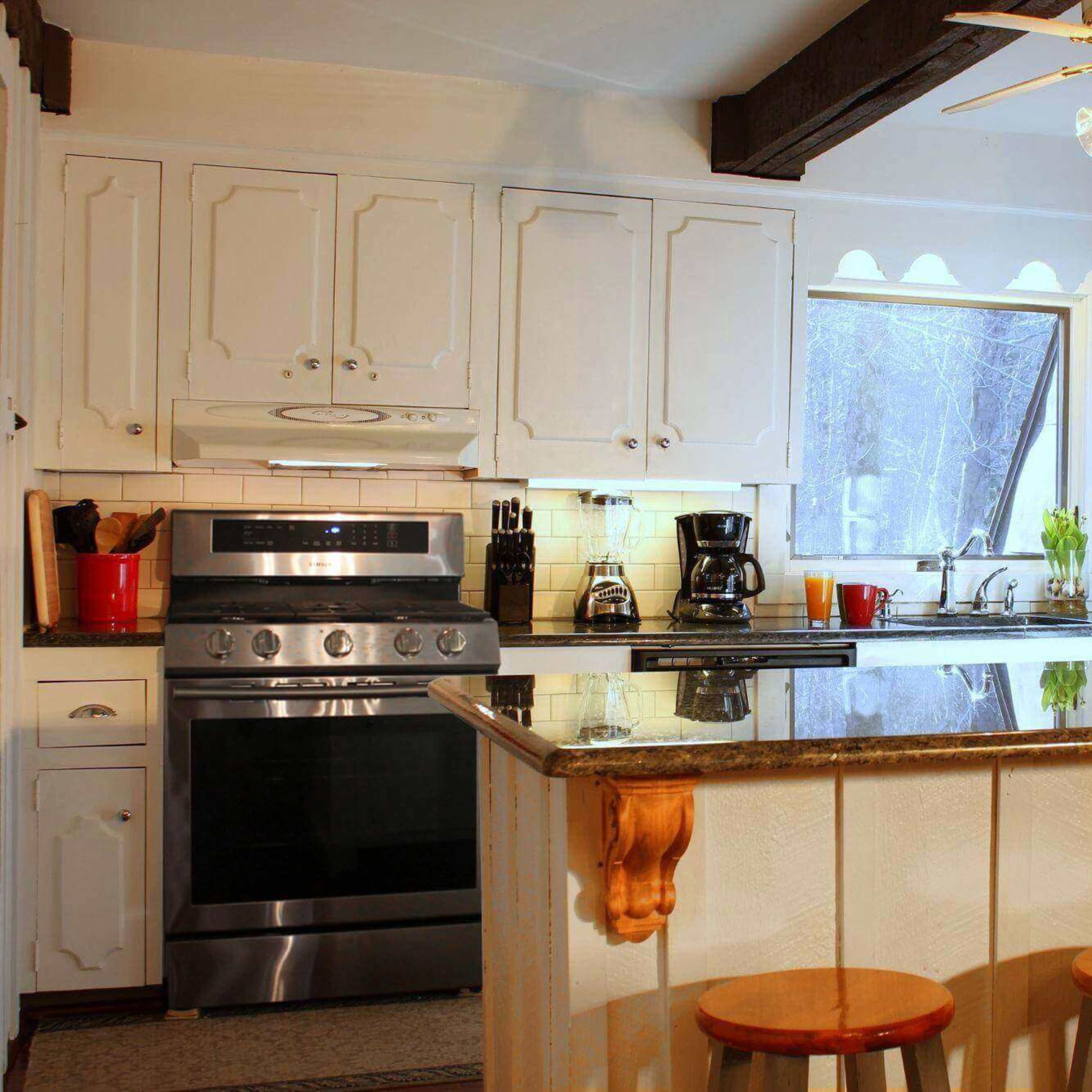 A modern kitchen features white cabinetry, a large granite island with three wooden stools, and stainless steel appliances.