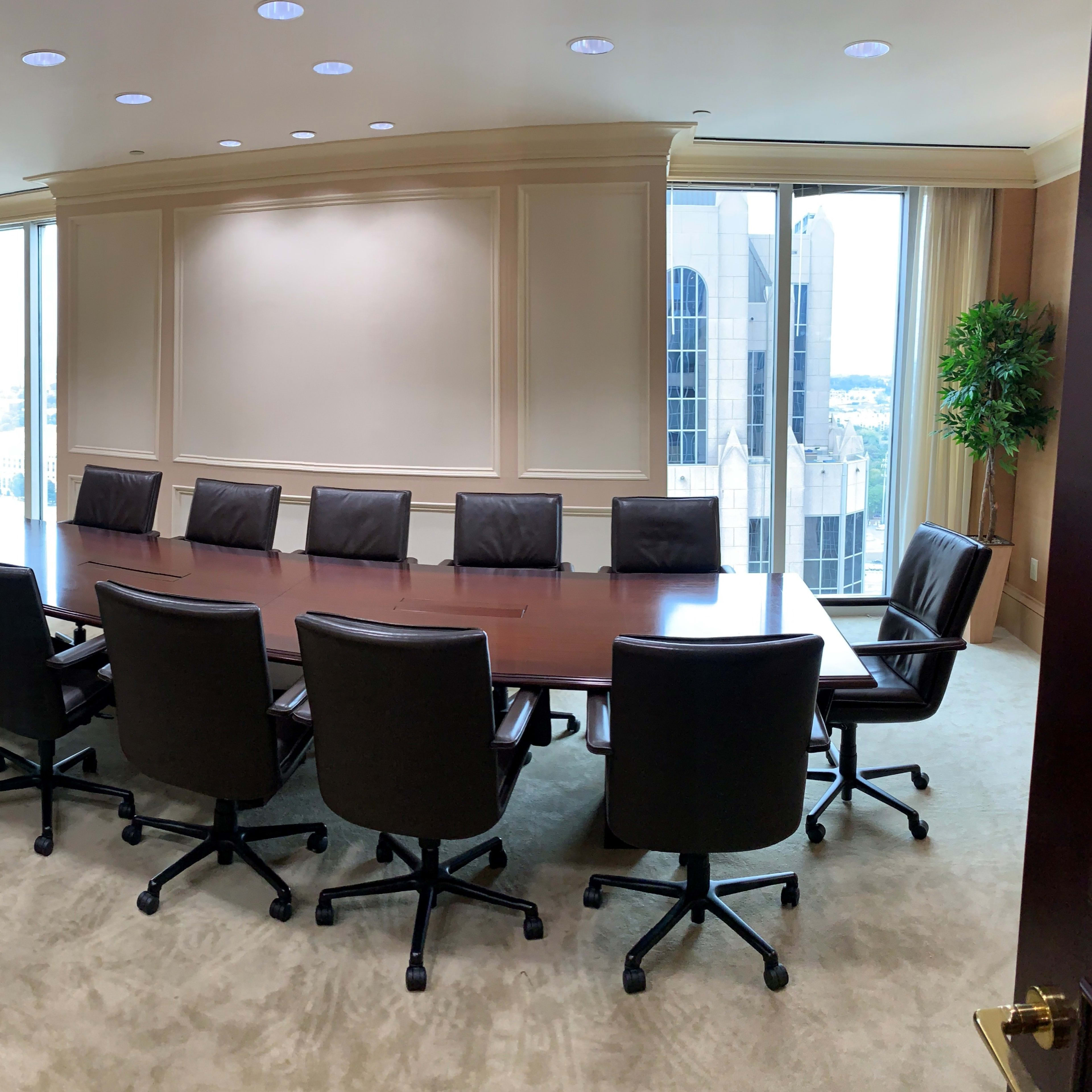 A conference room features a long wooden table surrounded by black leather chairs, with large windows displaying a city skyline.