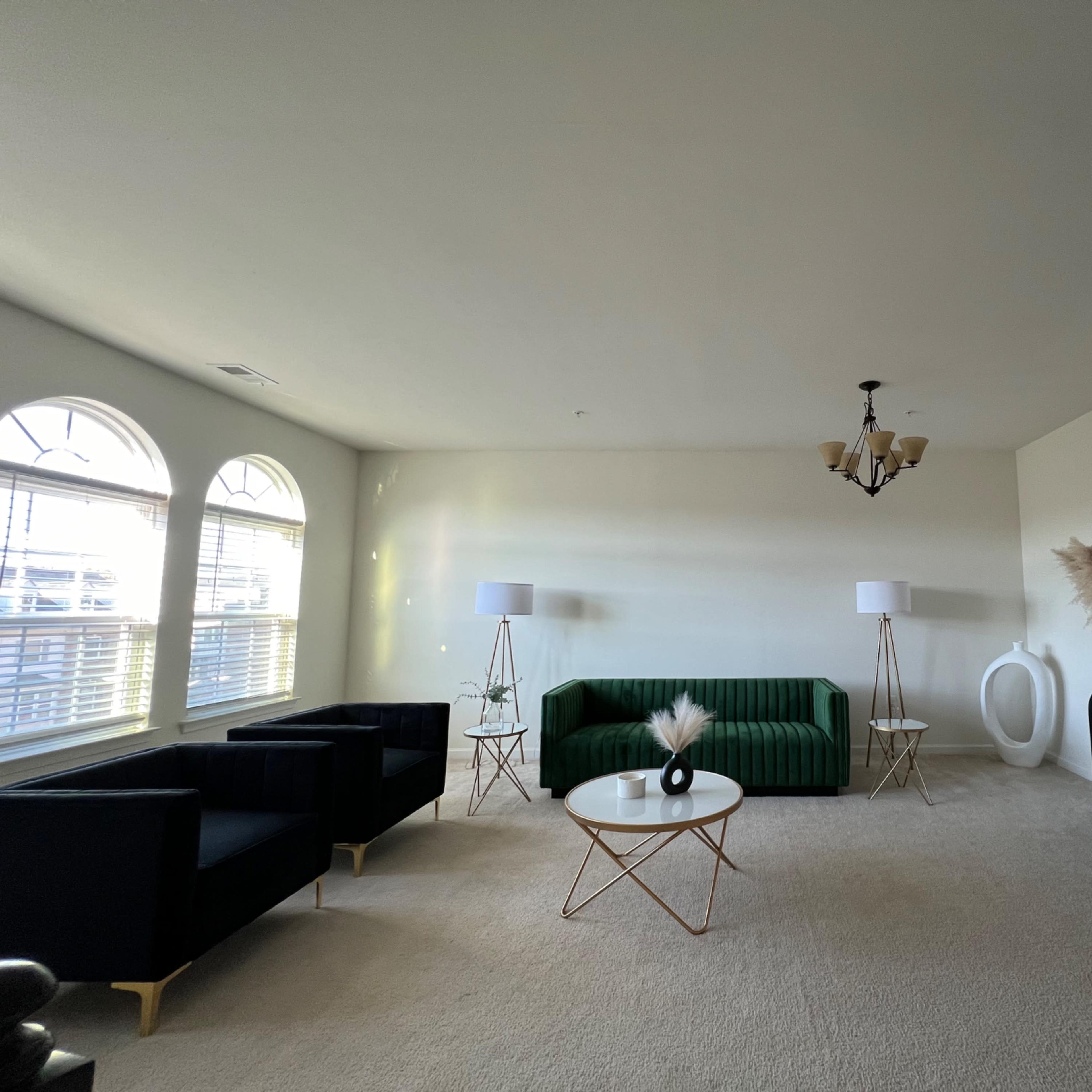 A spacious living room features a green sofa, two black armchairs, a round coffee table, and a console table adorned with decorative elements, under a chandelier and natural light from large windows.