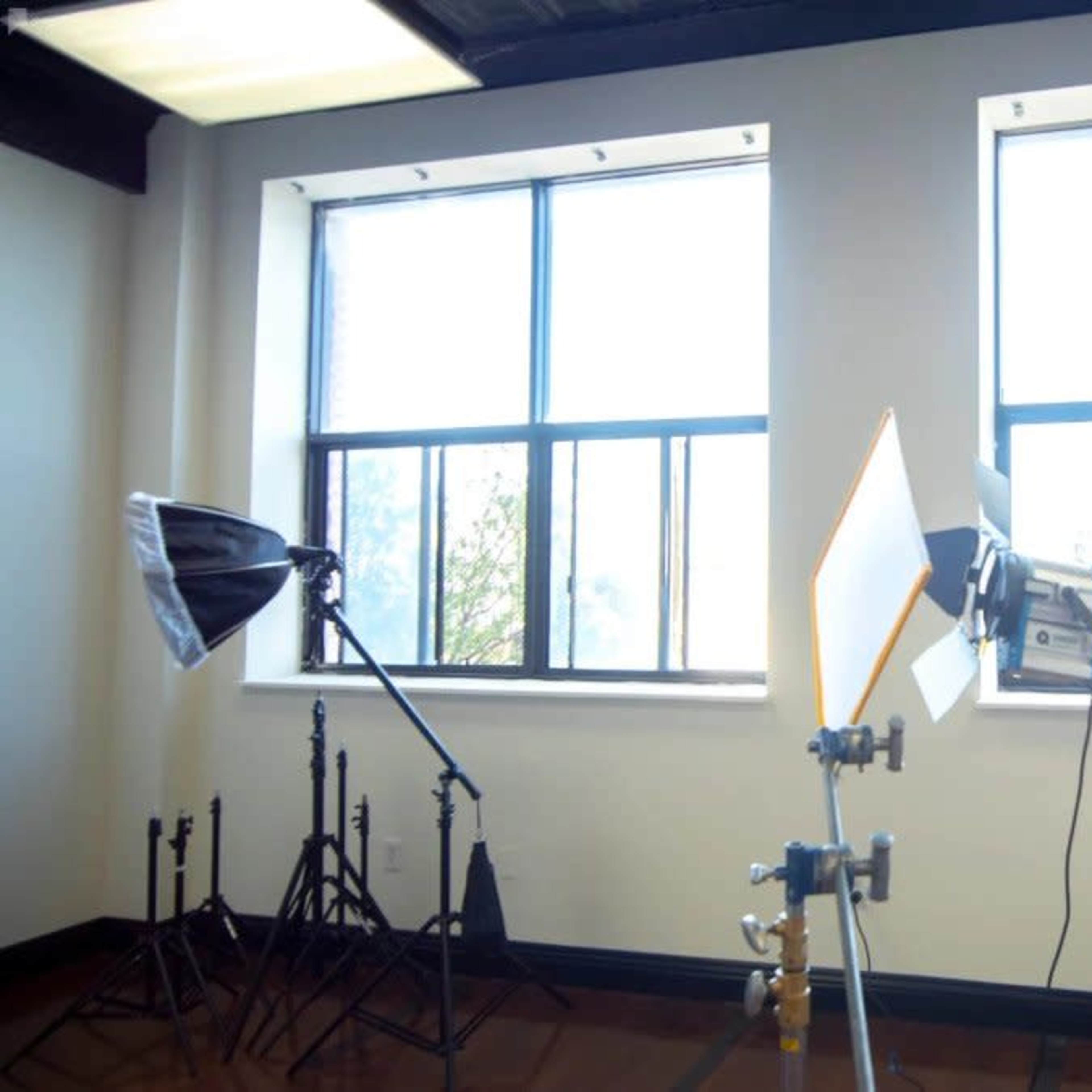 A well-lit studio space with two large windows and several lighting equipment setups positioned in front of them.