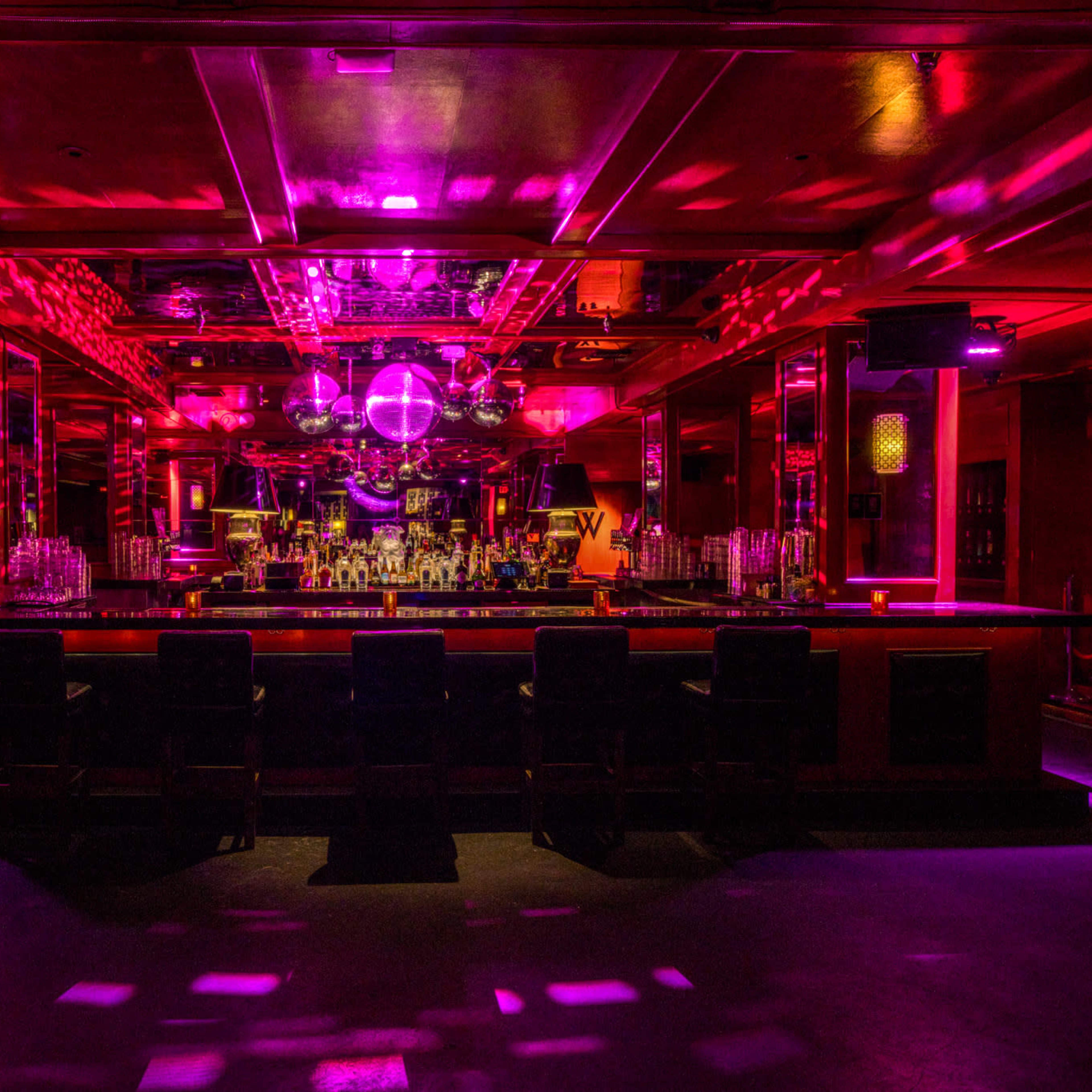 A dimly lit bar features a long counter with empty stools, illuminated by vibrant purple and pink lights and adorned with disco balls overhead.