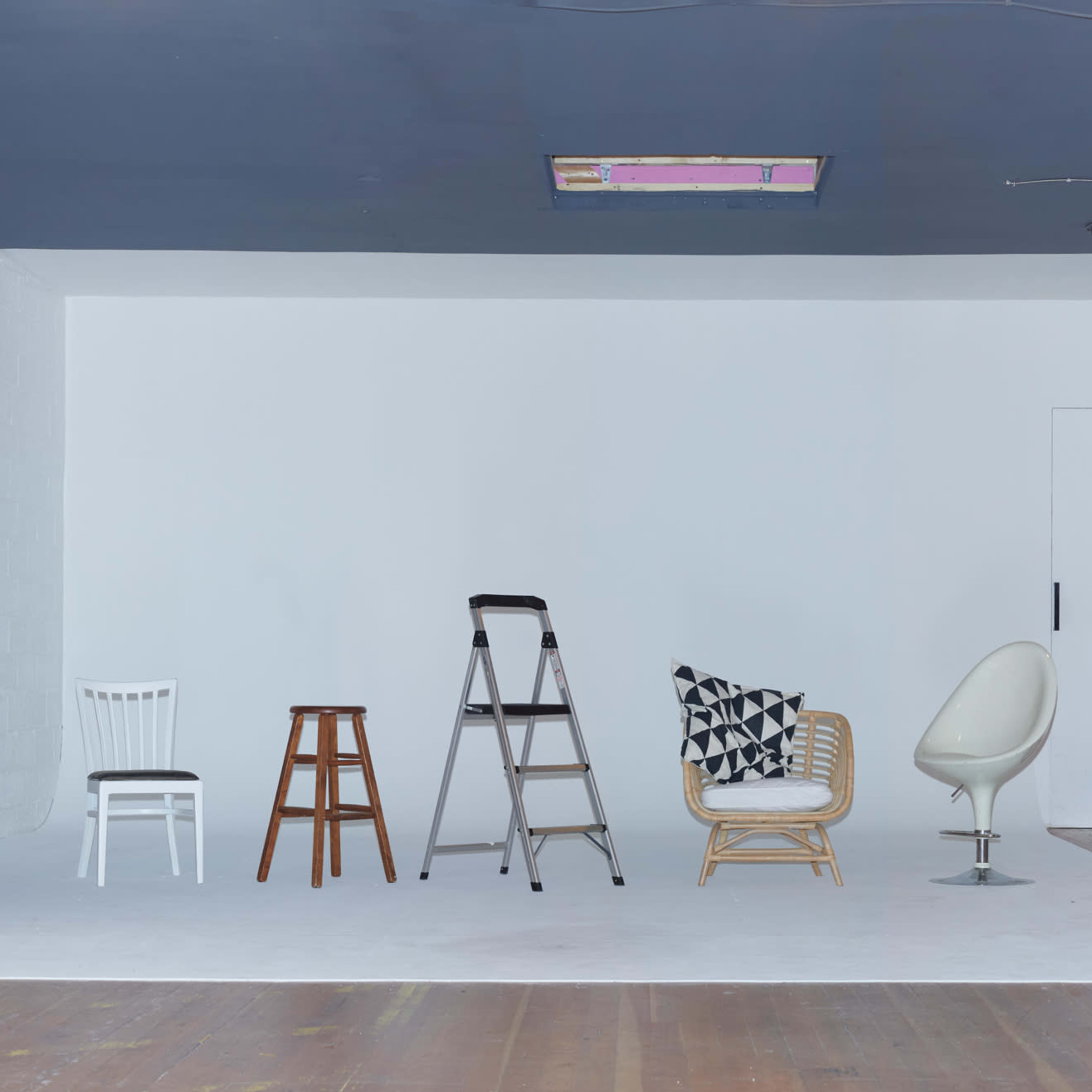 A spacious photography studio with a wooden floor, white backdrop, and several chairs and stools arranged in the center.