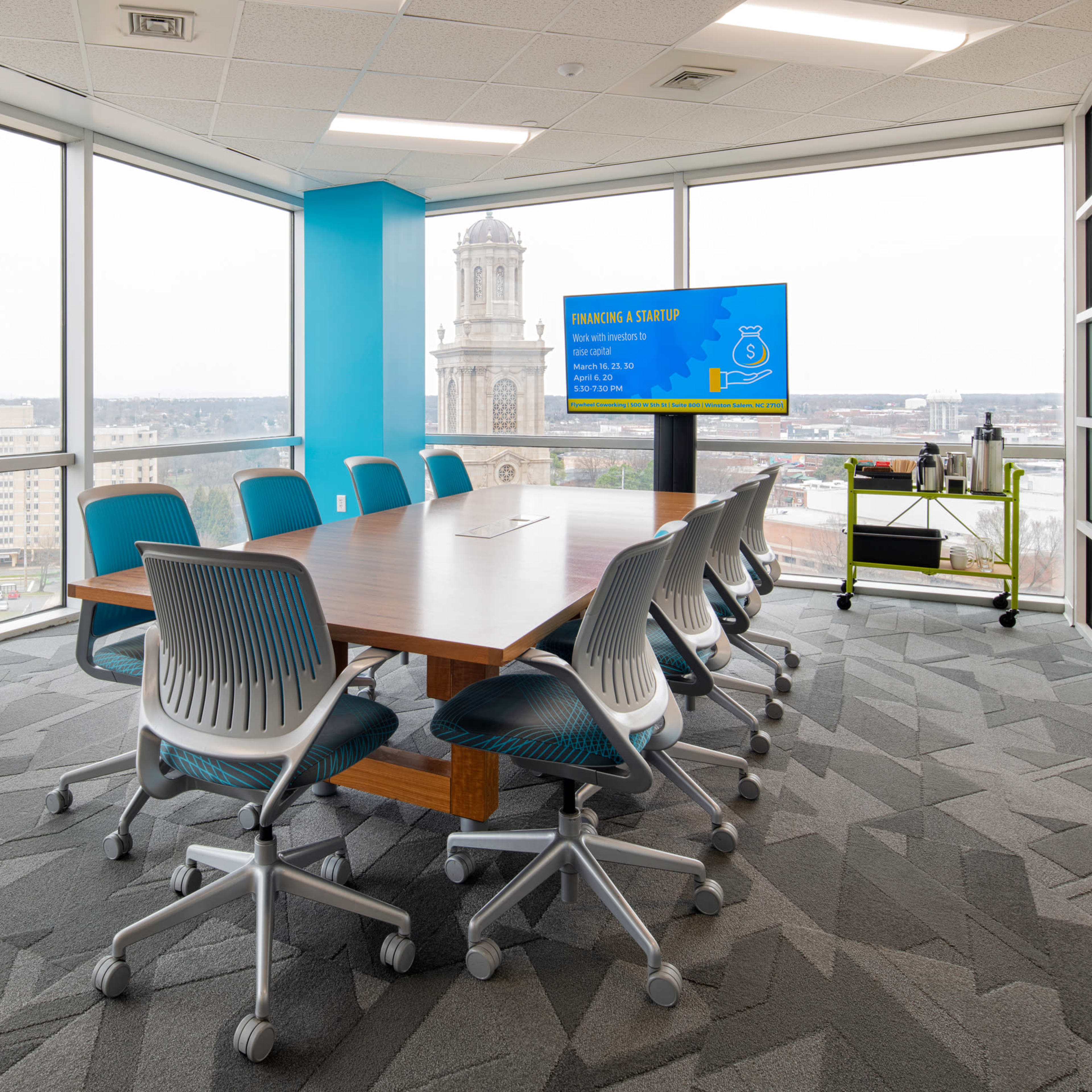 A modern conference room features a large wooden table surrounded by ergonomic chairs, with a screen displaying information and whiteboards for brainstorming, all in a sunlit space with views of the city outside.