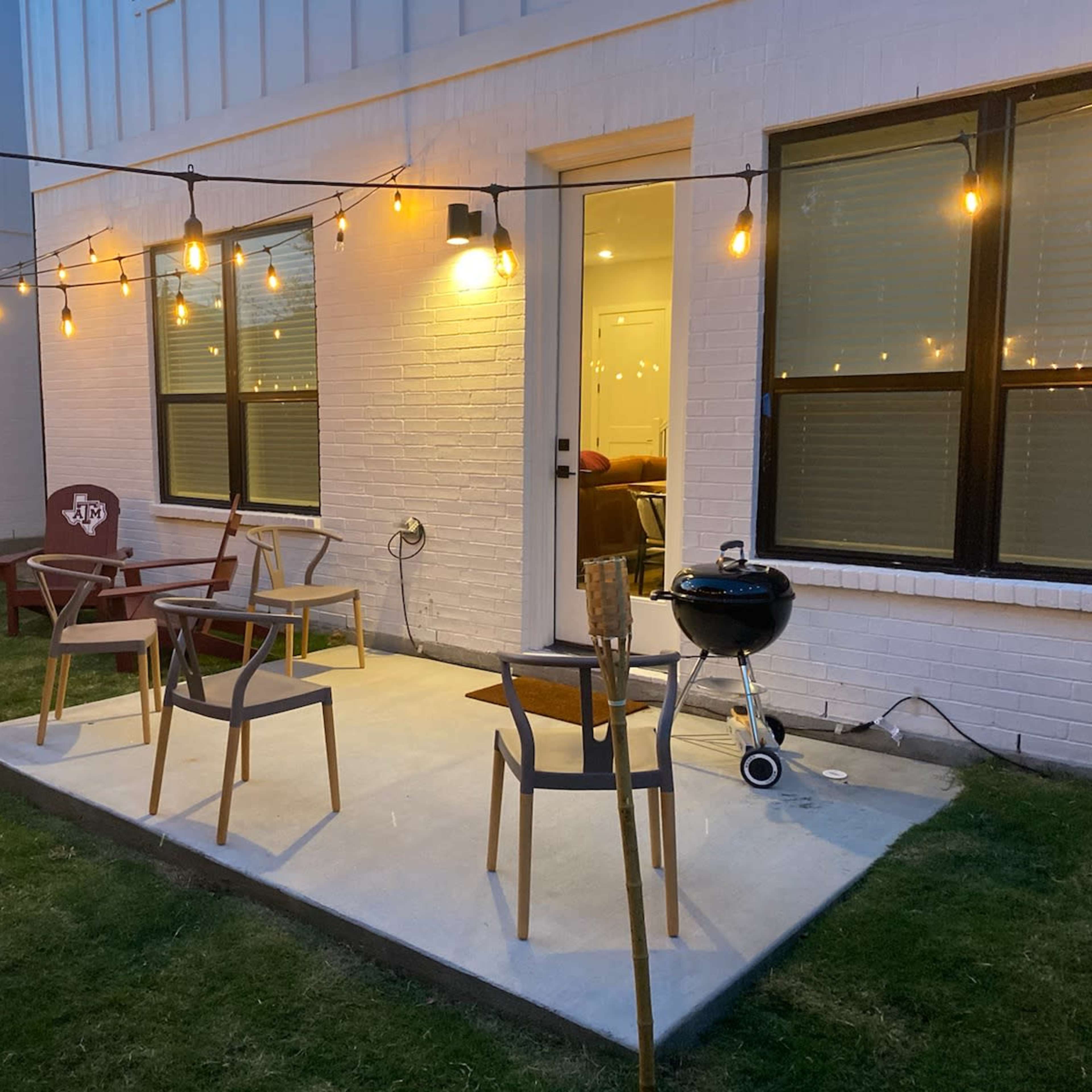 A patio area with several chairs and a barbecue grill, illuminated by string lights above.