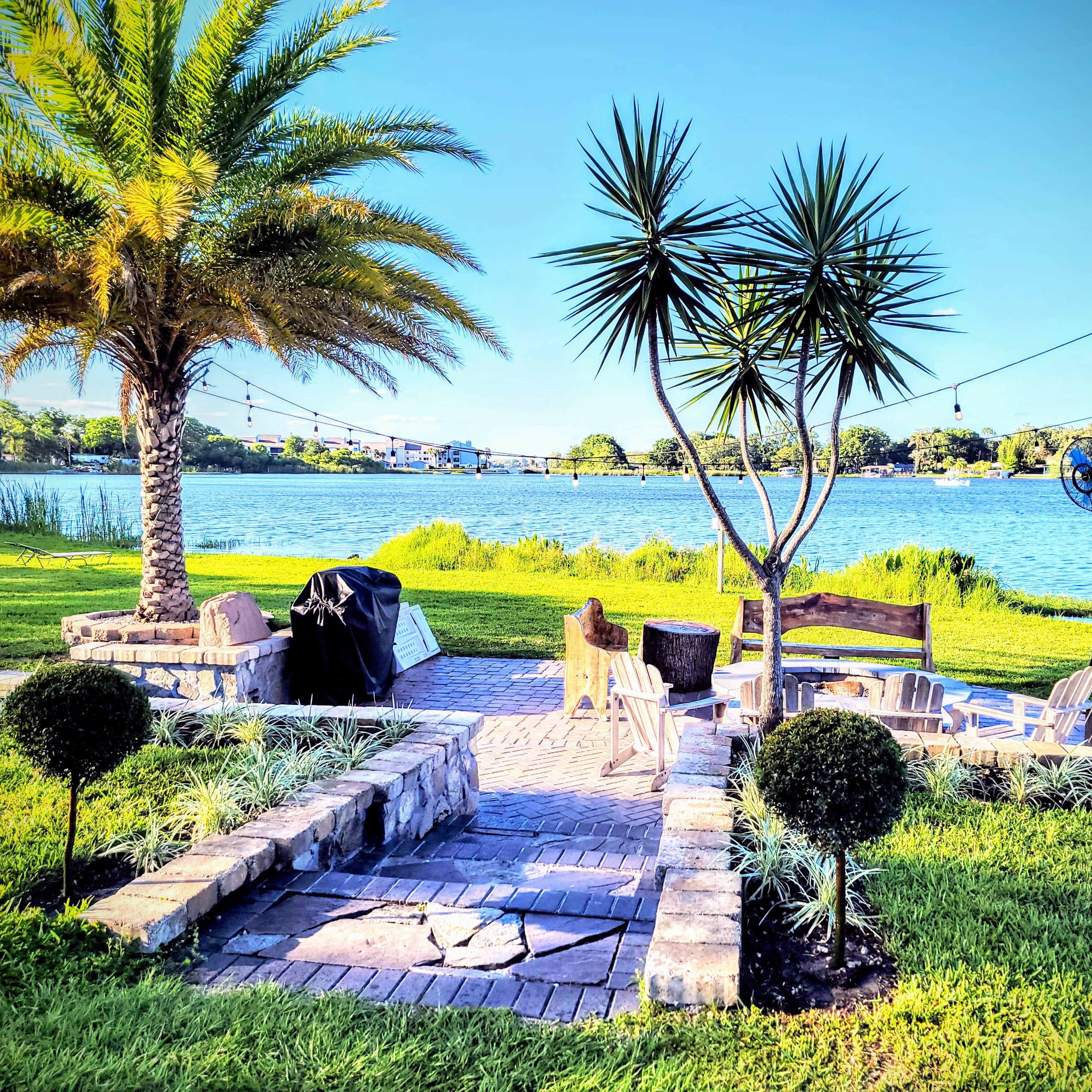 A landscaped yard by a lake, featuring a seating area with chairs and a grill, surrounded by palm trees and decorative plants.
