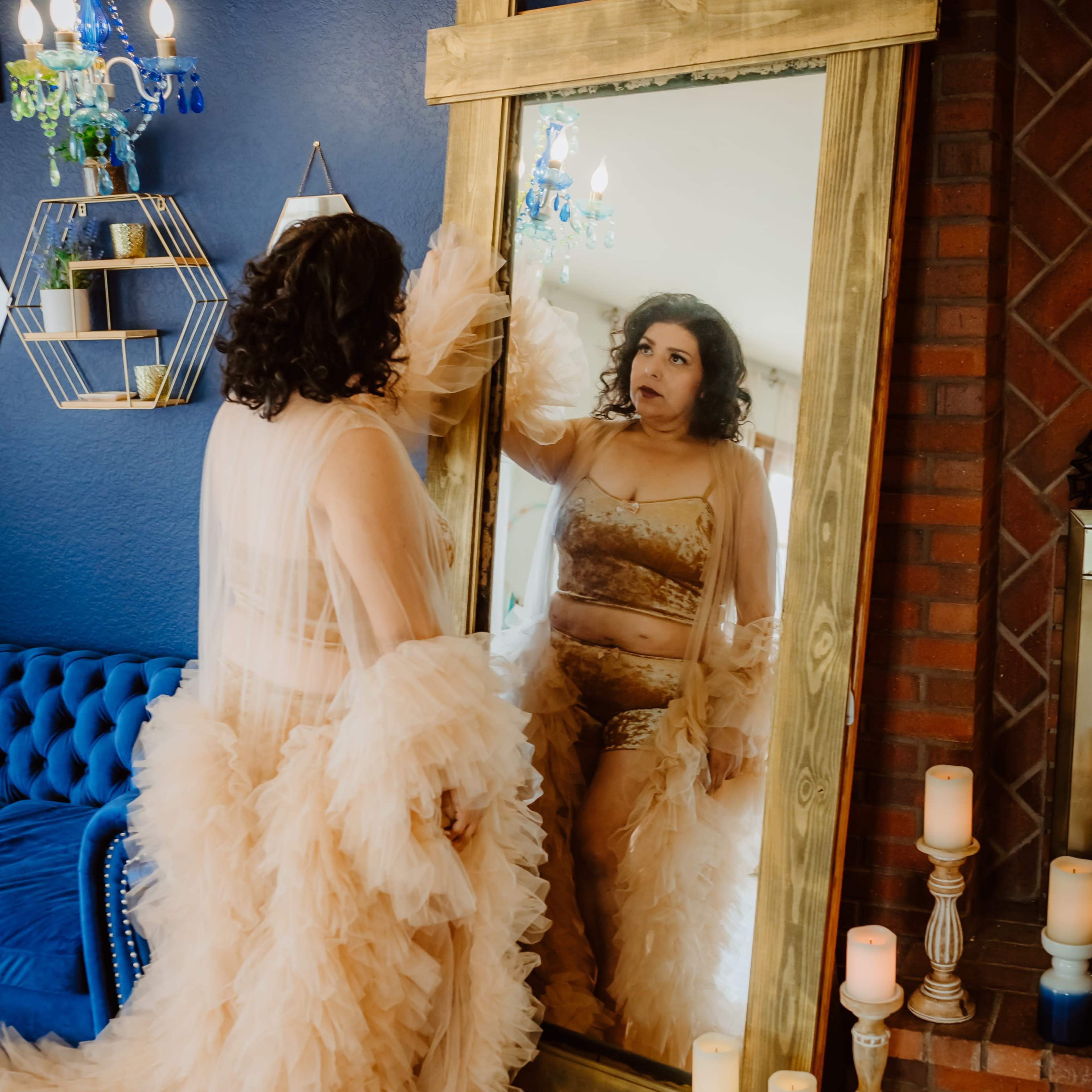 A woman in a flowing, layered gown stands in front of a large mirror, examining her reflection in a stylishly decorated room with a blue velvet sofa and candles.