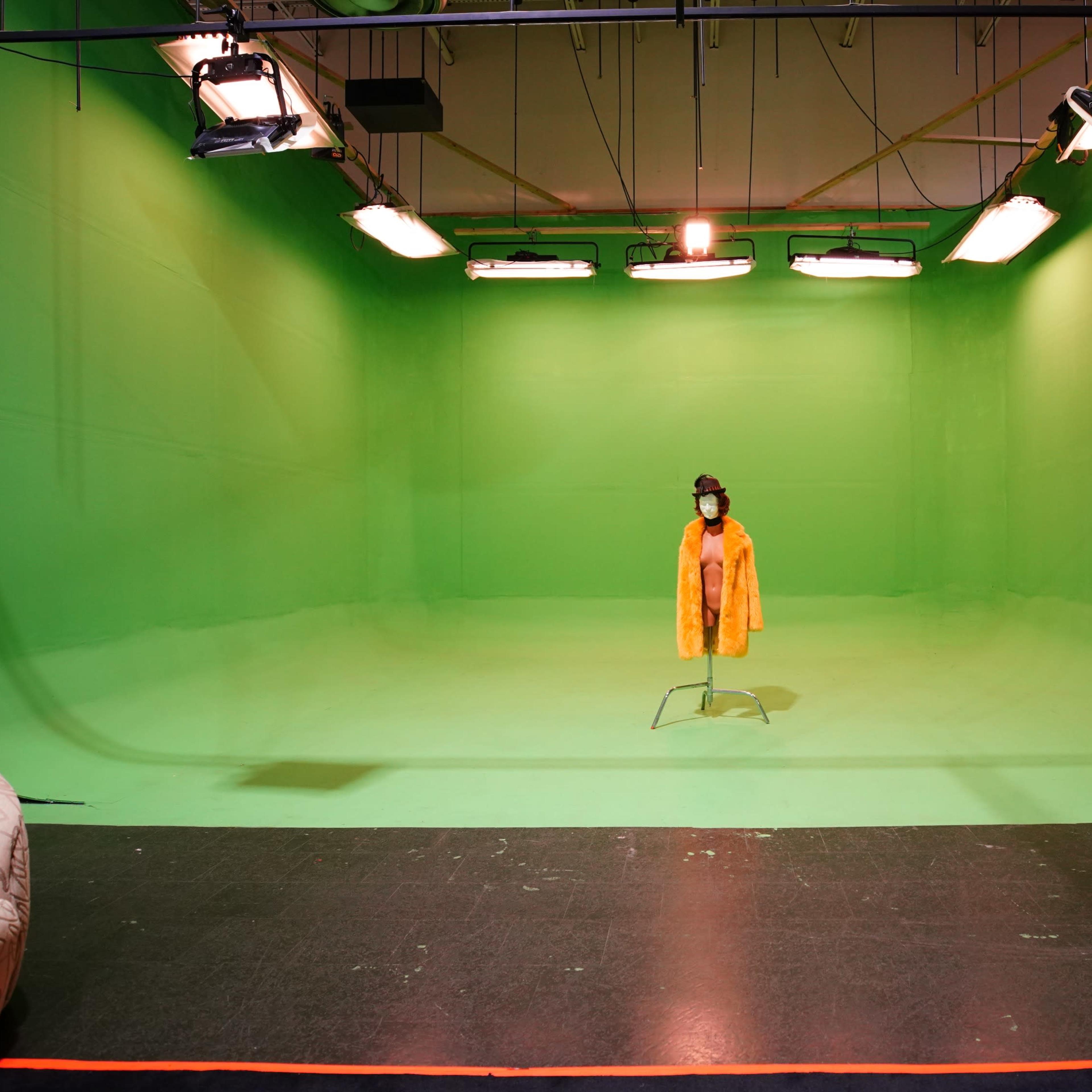 A figure in a yellow coat stands alone on a set with a green screen backdrop and studio lighting.