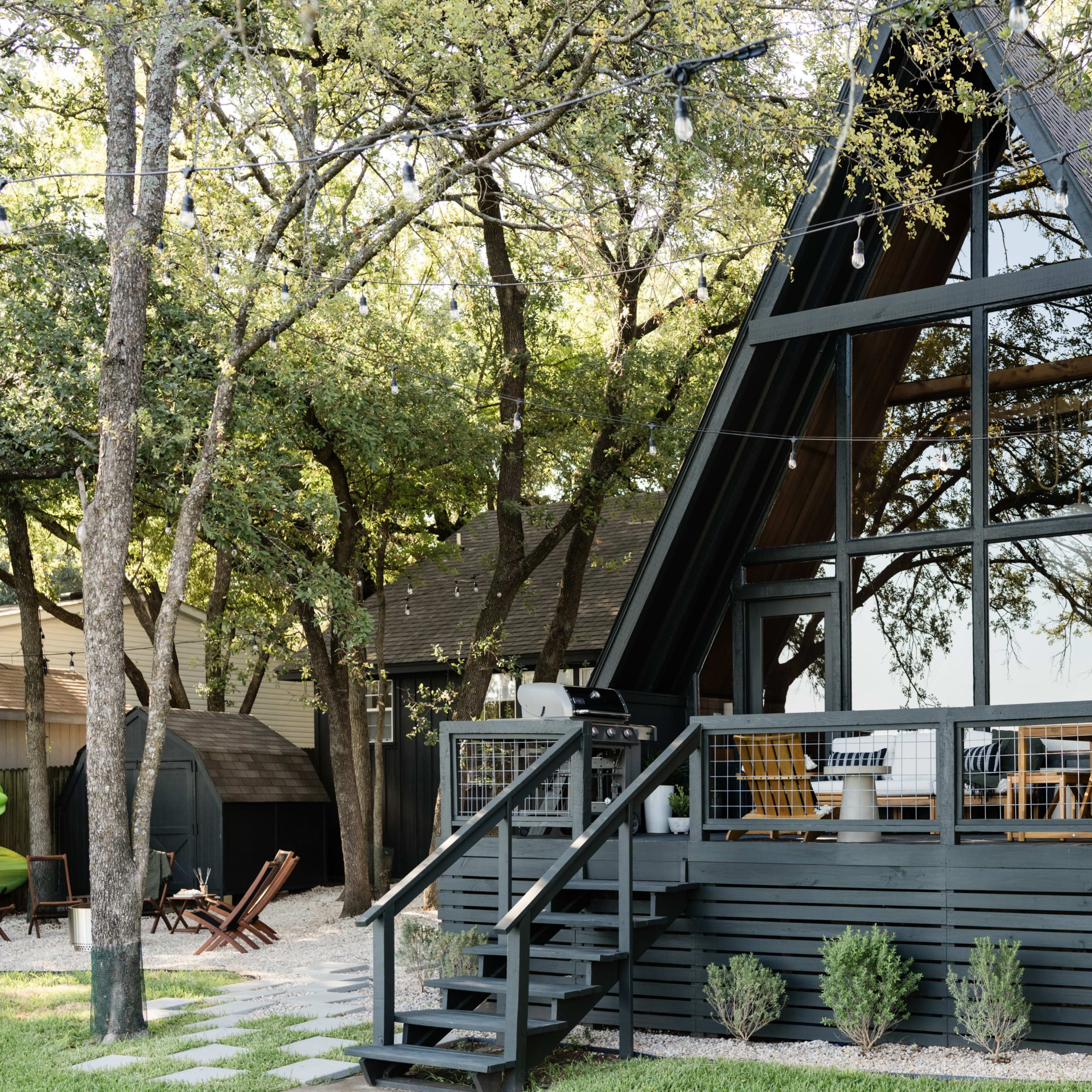 A black A-frame cabin with large windows is surrounded by trees and features a deck with outdoor seating.