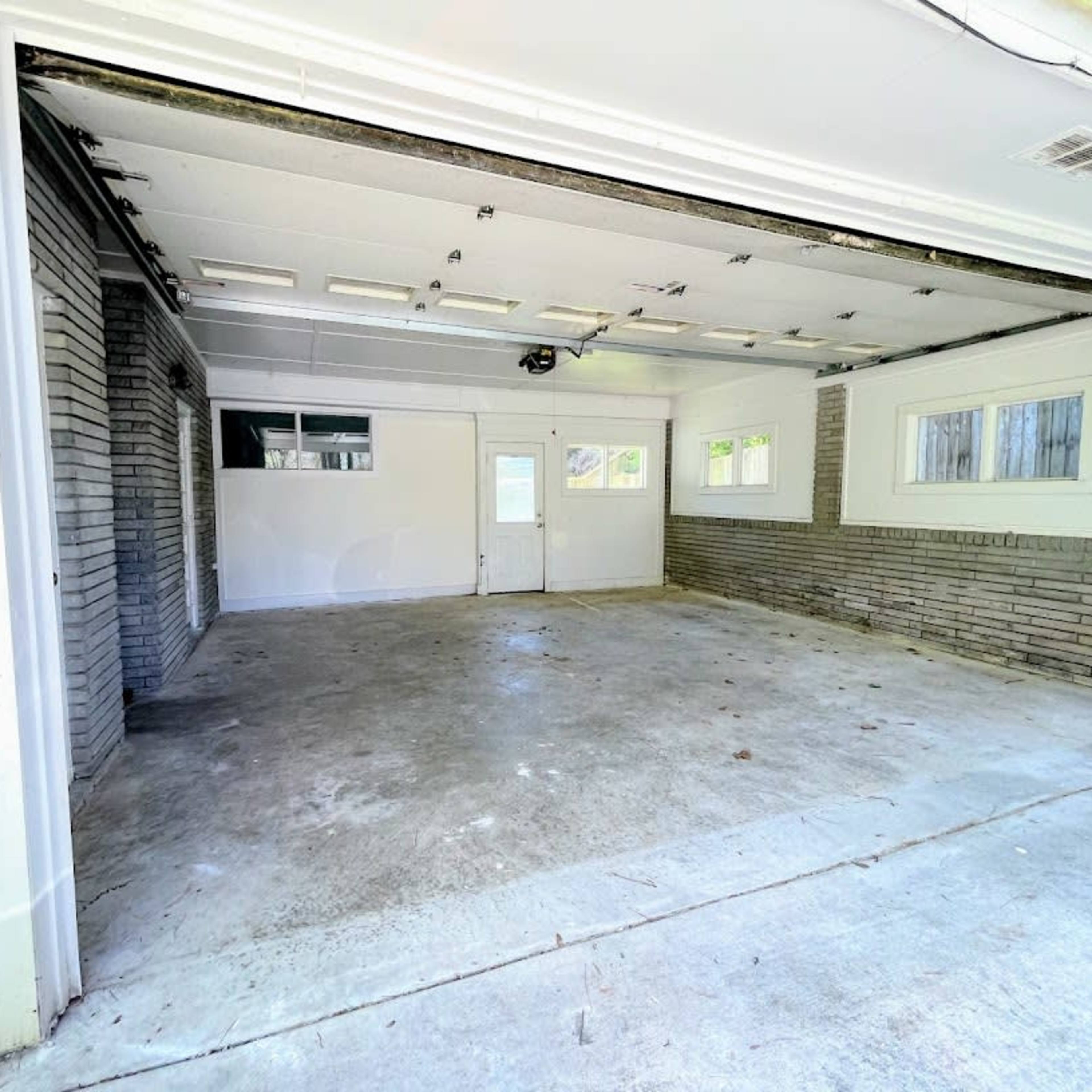 An empty garage with concrete flooring and block walls, featuring large windows and a door at the far end.