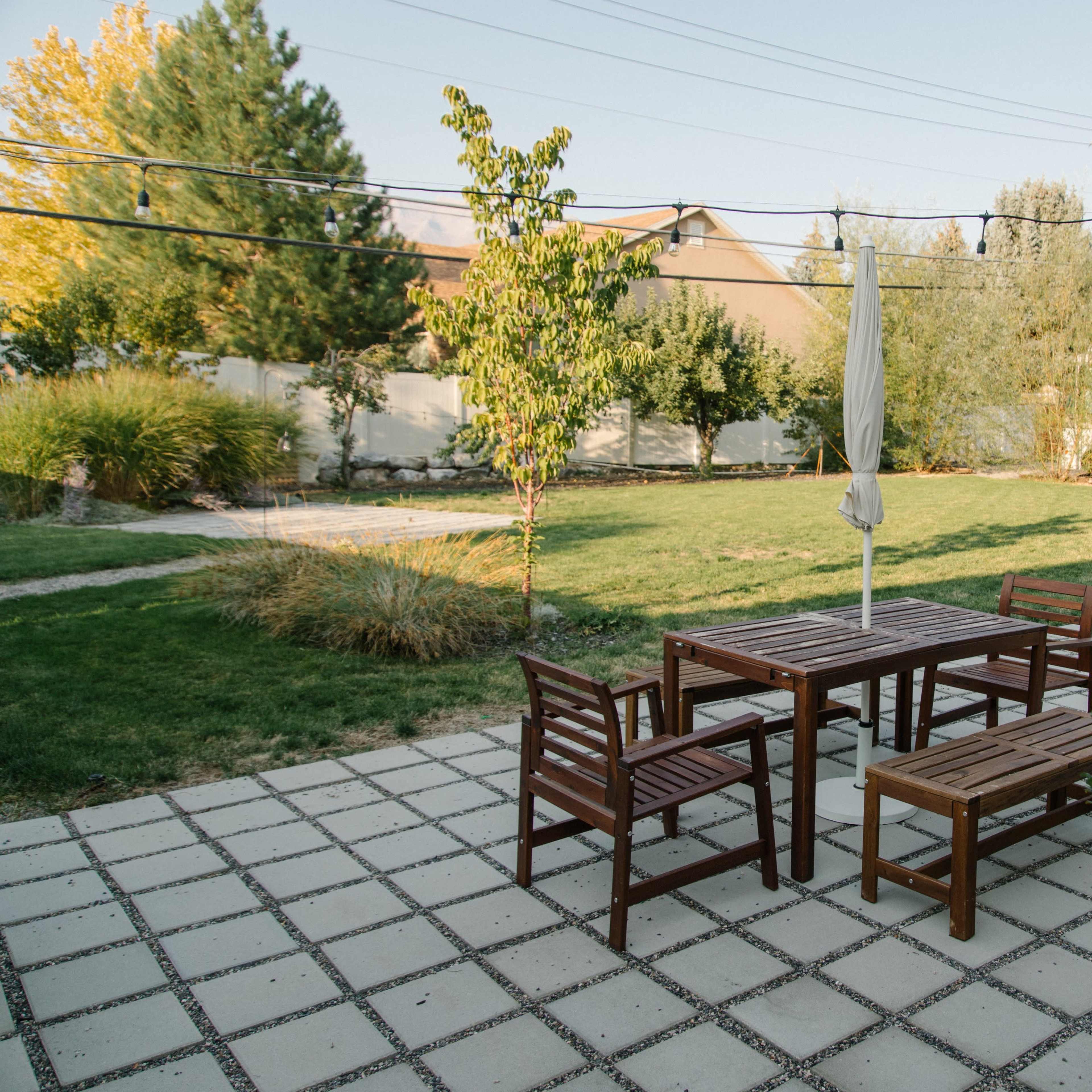 A patio with wooden furniture, including a table, chairs, and a bench, set against a grassy yard and trees.
