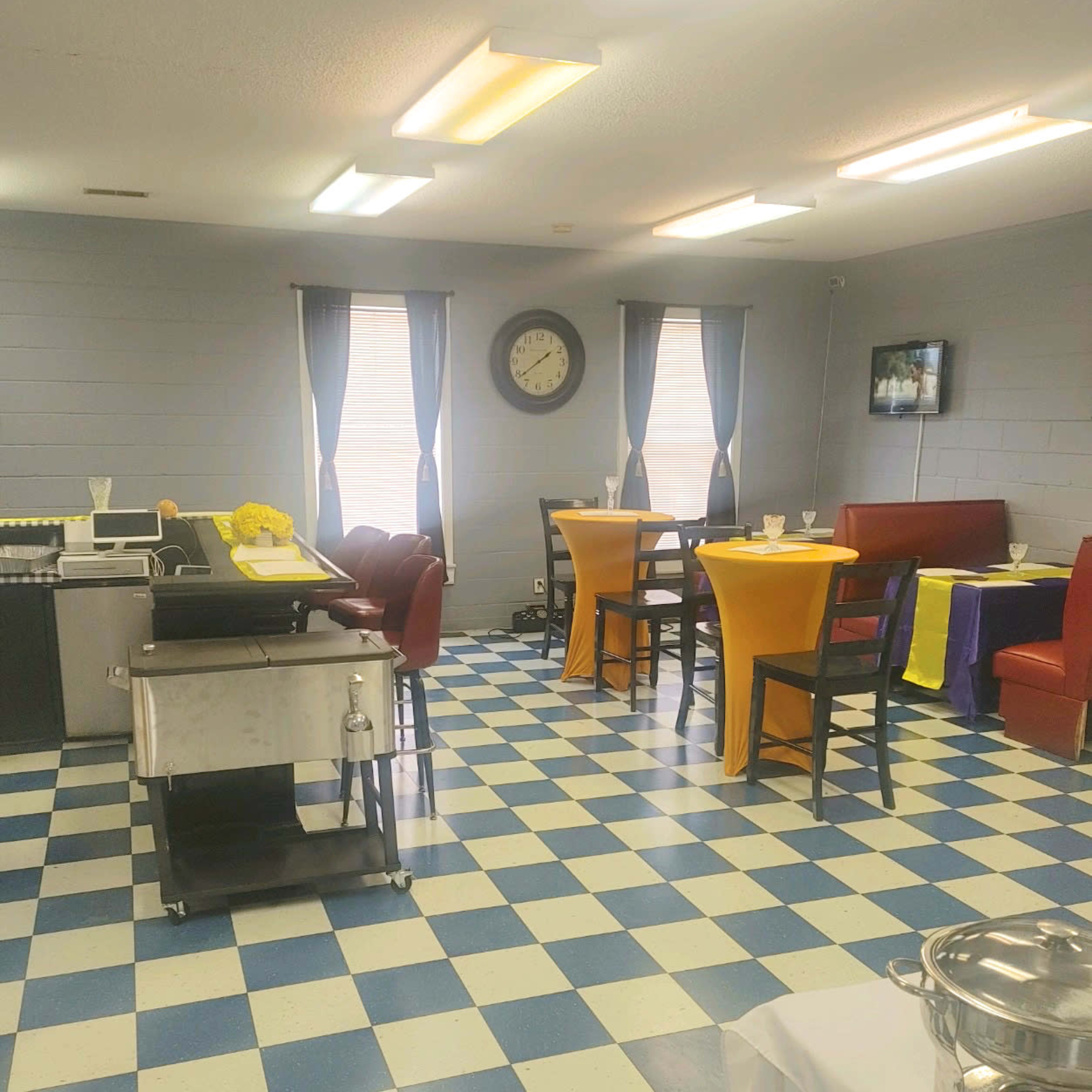 A dining area with a checkered blue and white floor, tables dressed in yellow and purple tablecloths, and a clock on the wall.