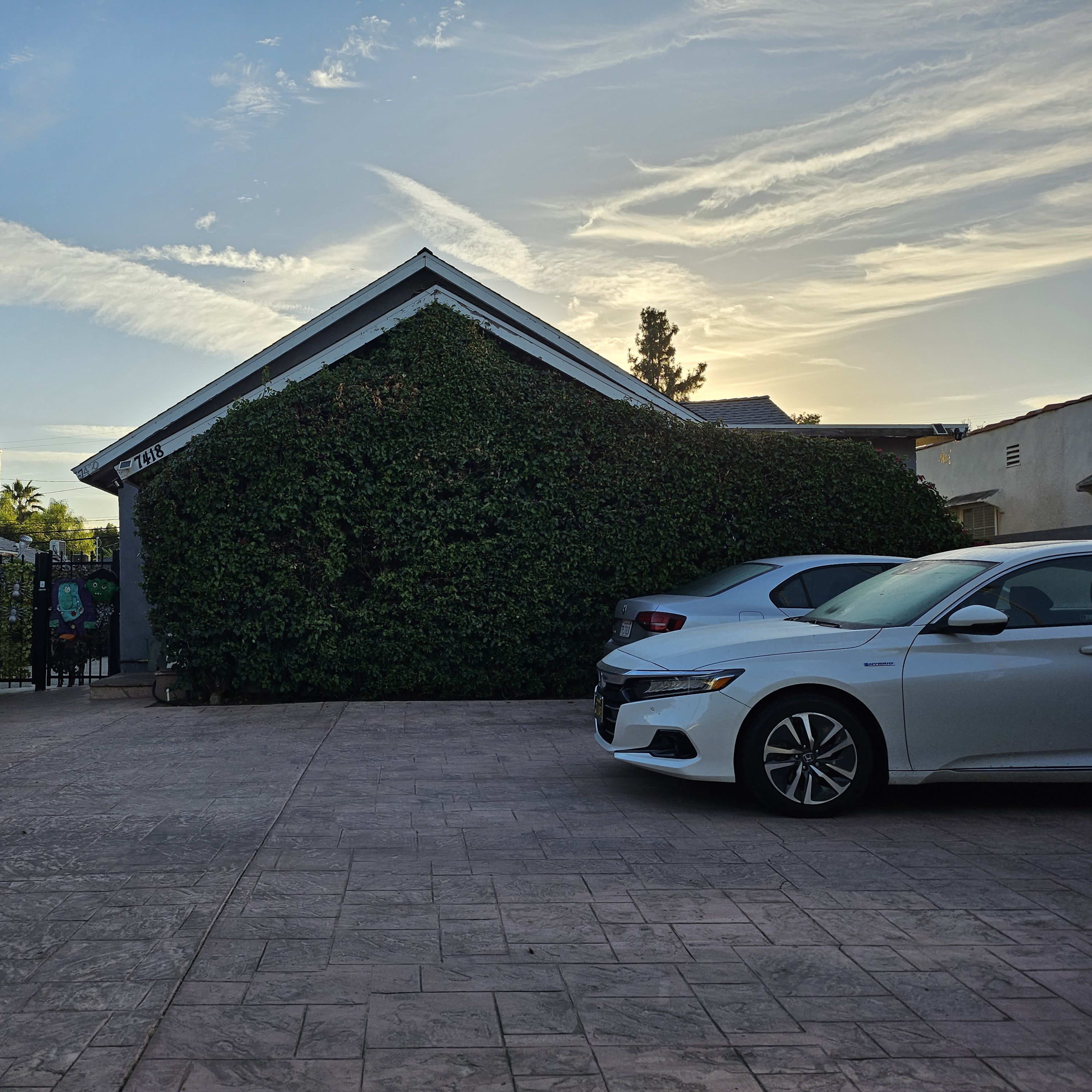 A house covered in greenery stands next to two parked cars in a paved driveway under a cloudy sky at sunset.