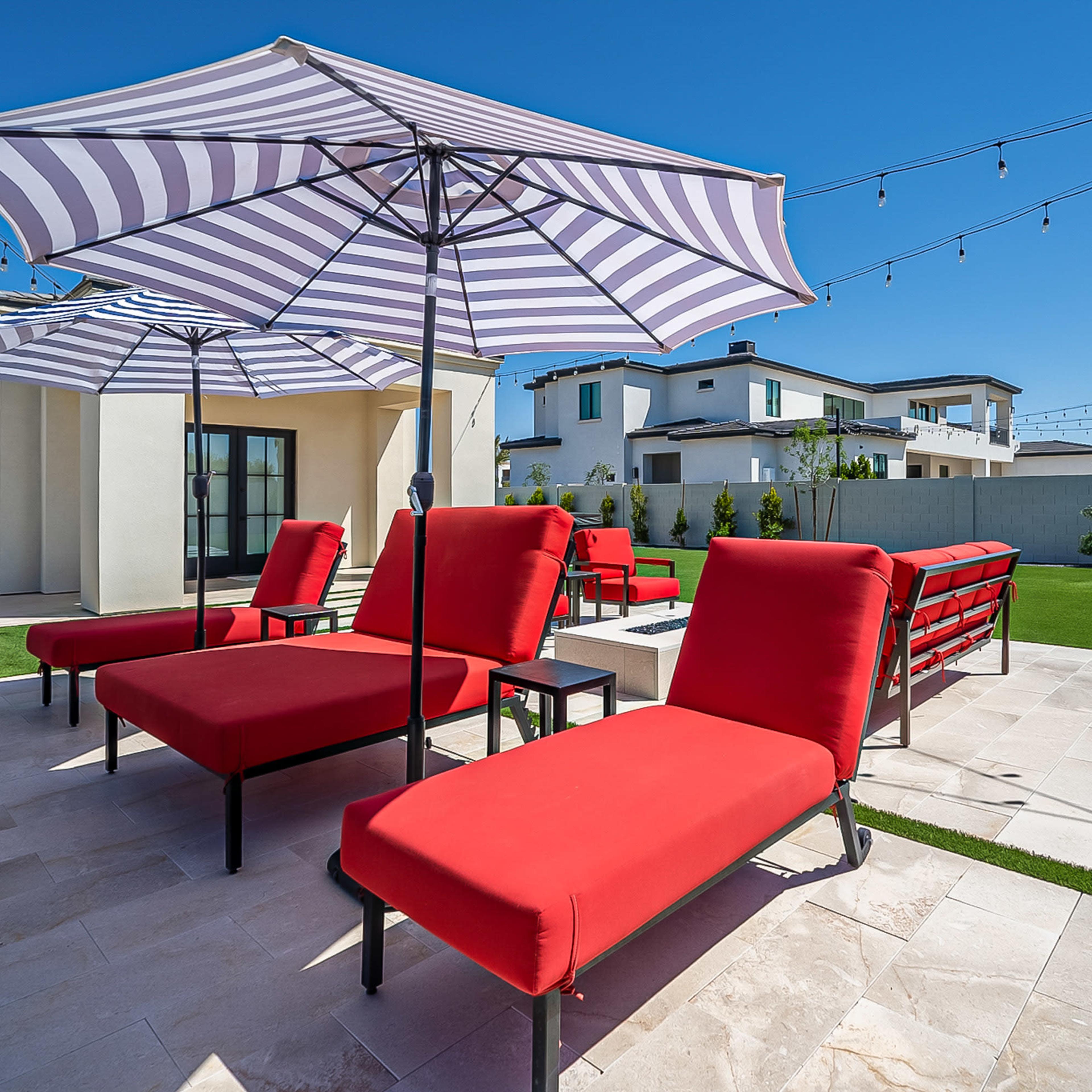 A landscaped outdoor area with red lounge chairs under striped umbrellas, set on a stone patio surrounded by greenery and string lights overhead.