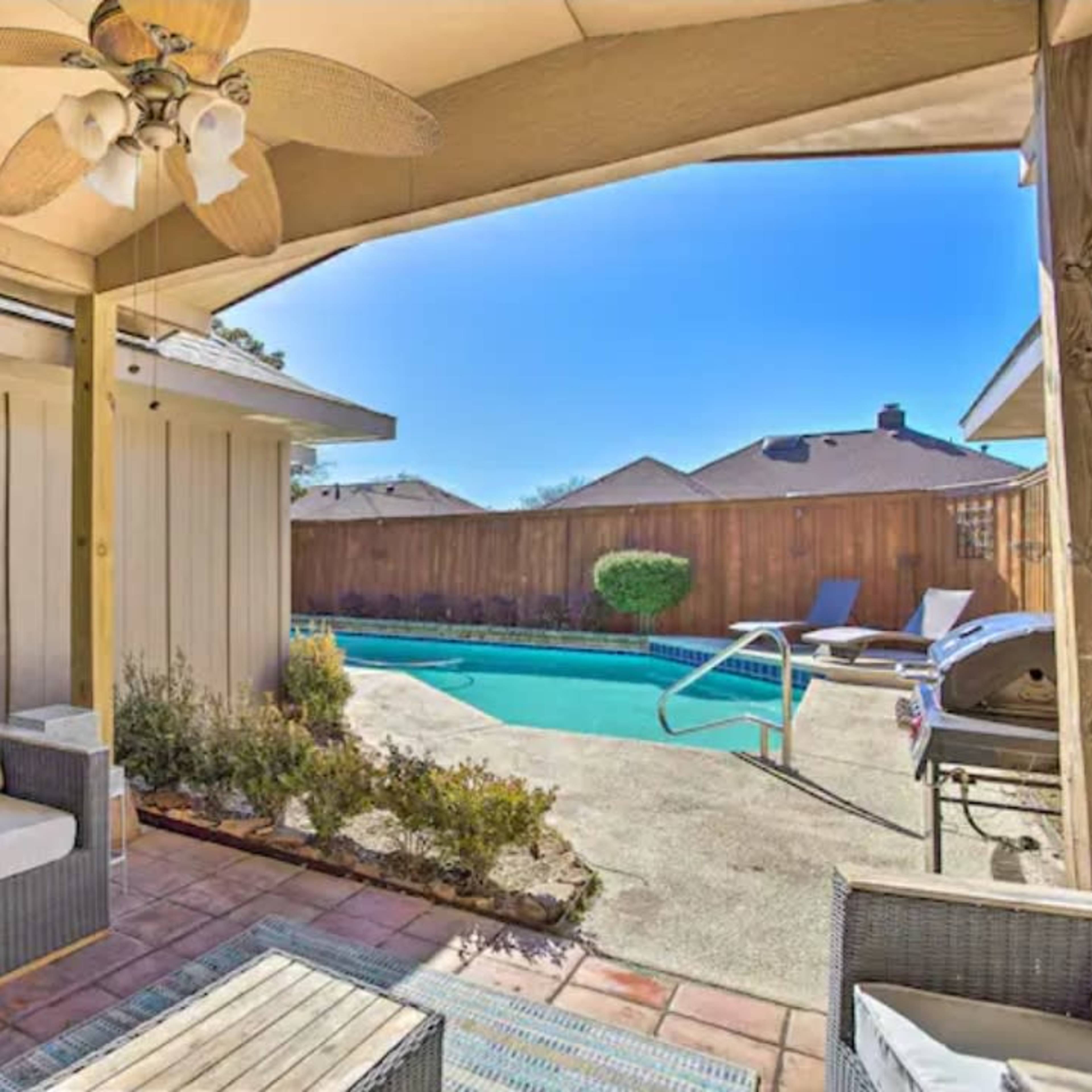A covered patio area with seating overlooks a swimming pool surrounded by a wooden fence.