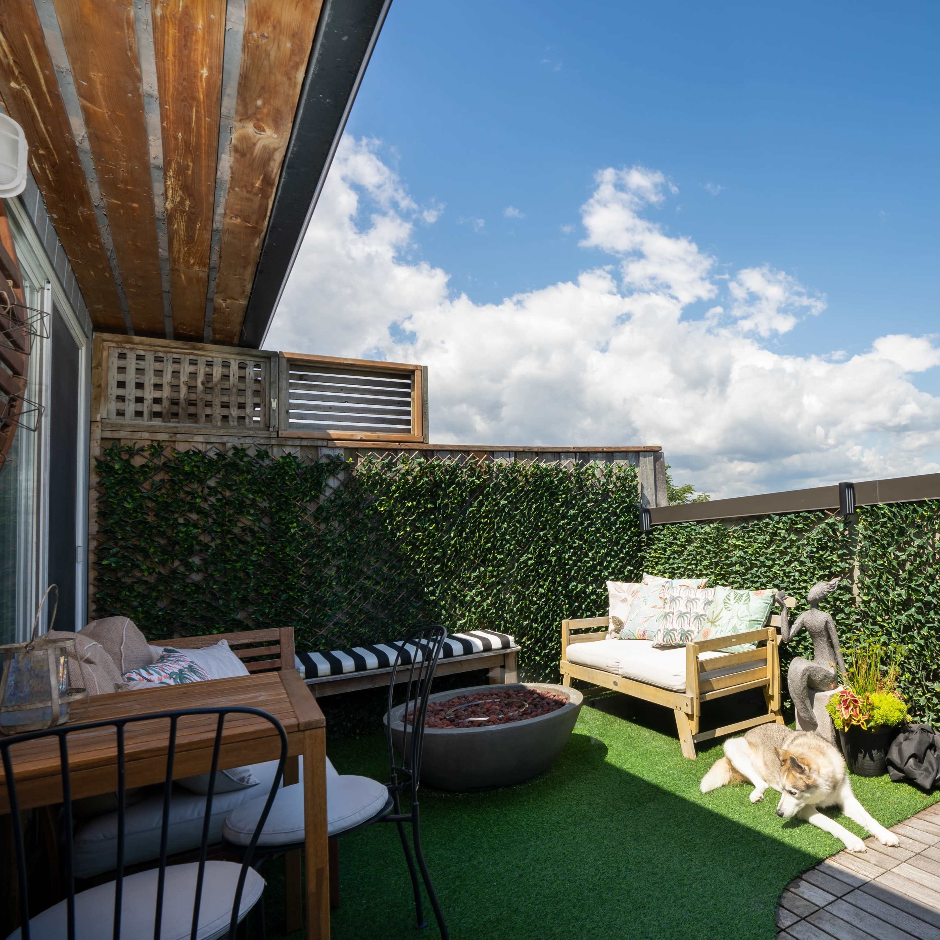 A balcony features a seating area with a sofa, a grill, and two dogs resting on artificial grass under a blue sky with clouds.