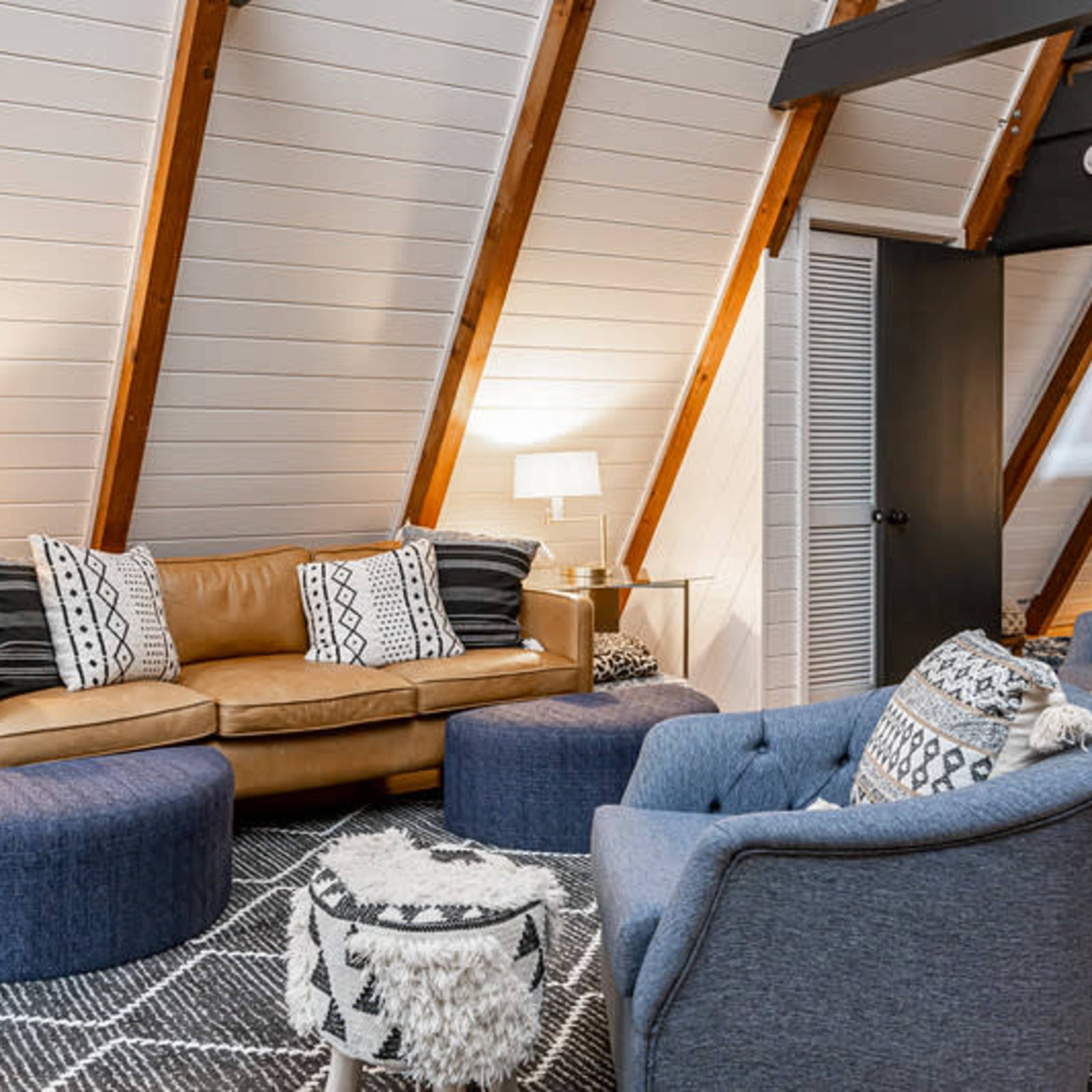 A cozy living room with sloped wooden ceilings, featuring a brown leather sofa, two blue ottomans, and a round coffee table on a patterned rug.