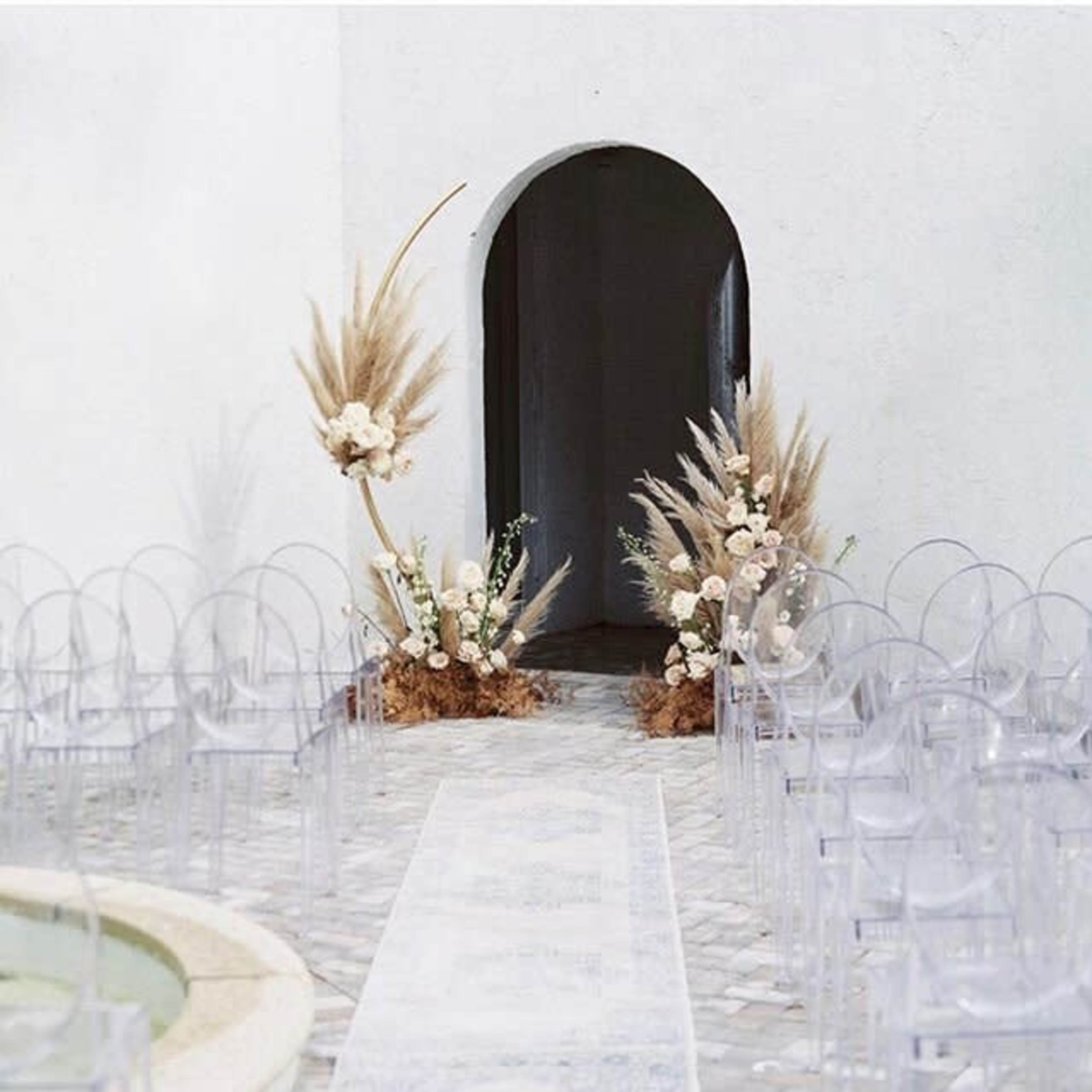 A wedding altar is set up with floral arrangements and transparent chairs lining a stone pathway leading to an arched entrance.