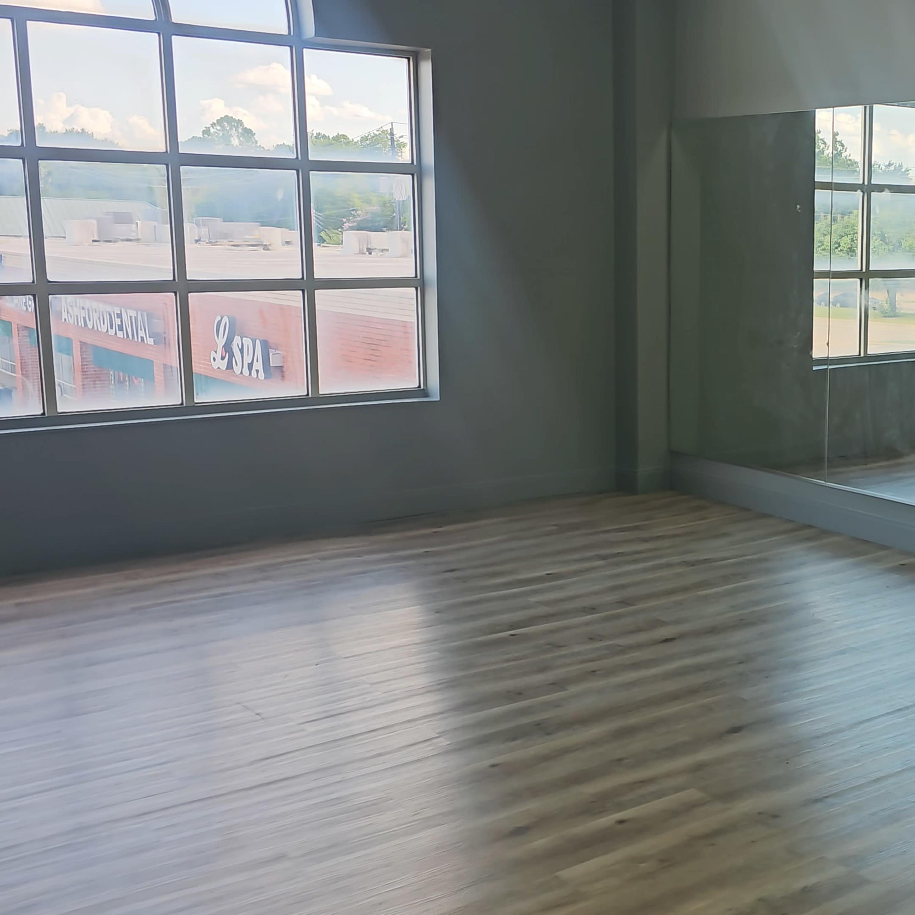A spacious studio with a wooden floor, large windows, and a mirrored wall, accompanied by a row of teal chairs.