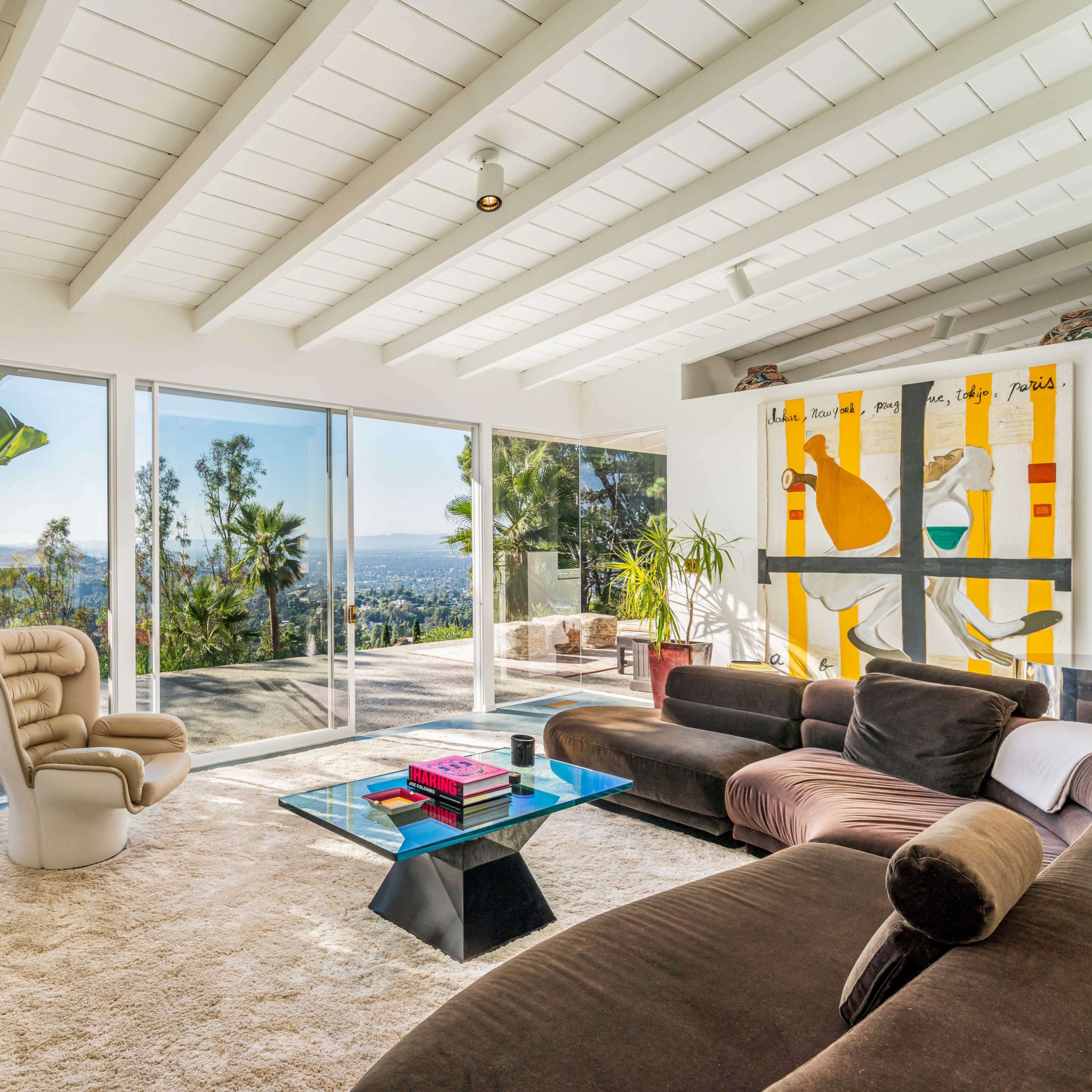 A spacious living room features large glass windows overlooking a scenic view, modern furniture, and vibrant artwork on the walls.