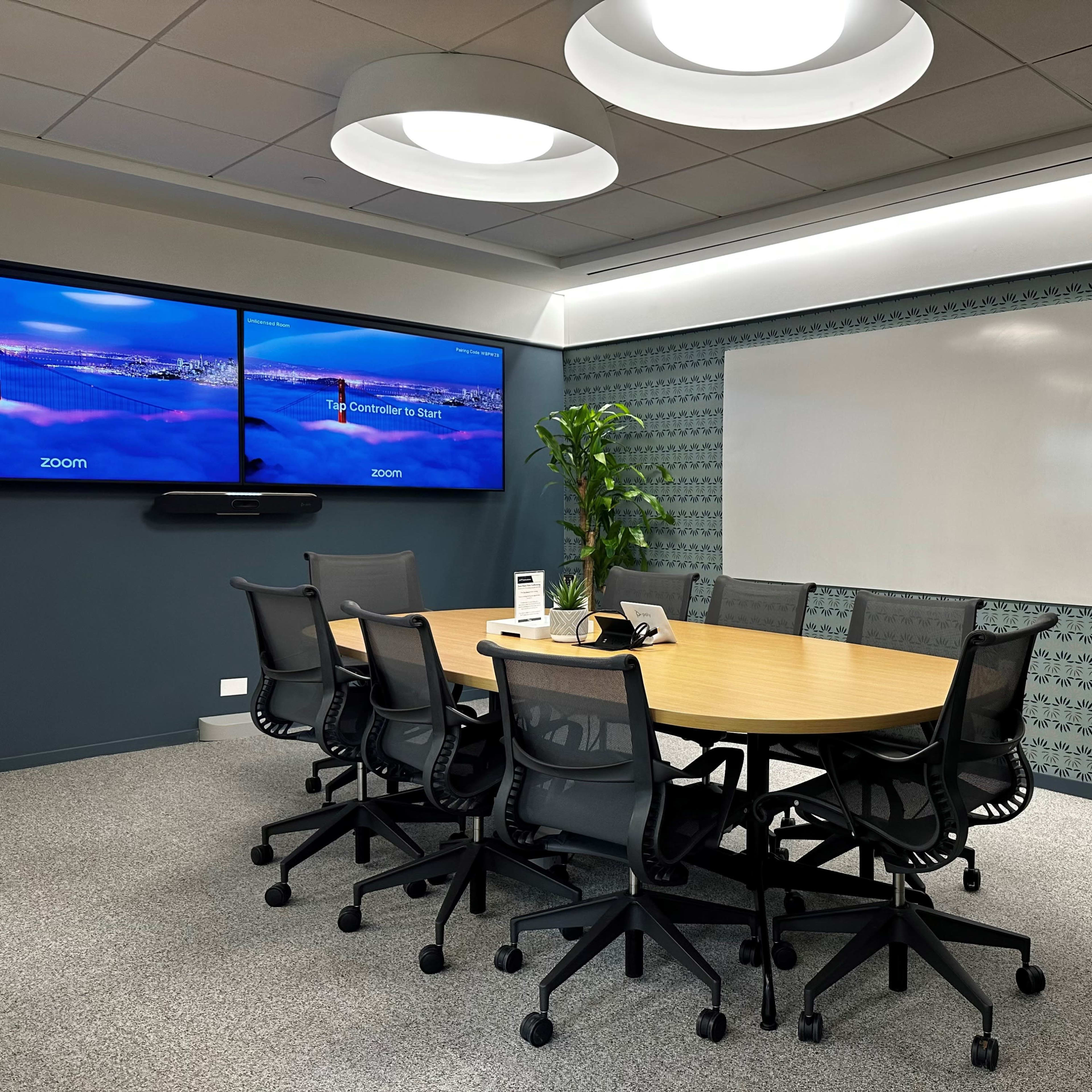 A modern conference room features a large wooden table surrounded by ergonomic chairs, two mounted screens displaying a Zoom interface, and a whiteboard on one wall.