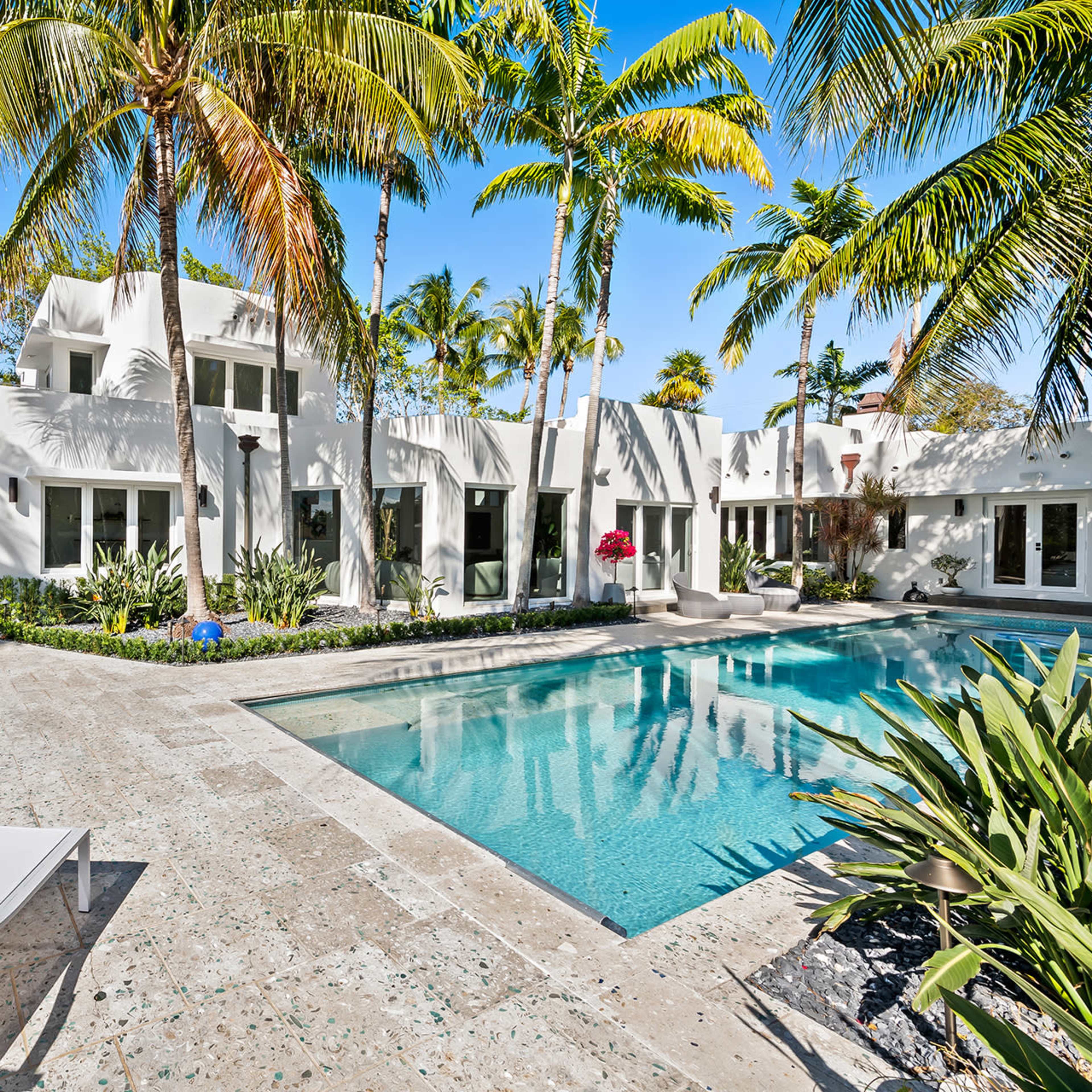 A modern white villa with large windows surrounds a swimming pool, framed by tropical palm trees.
