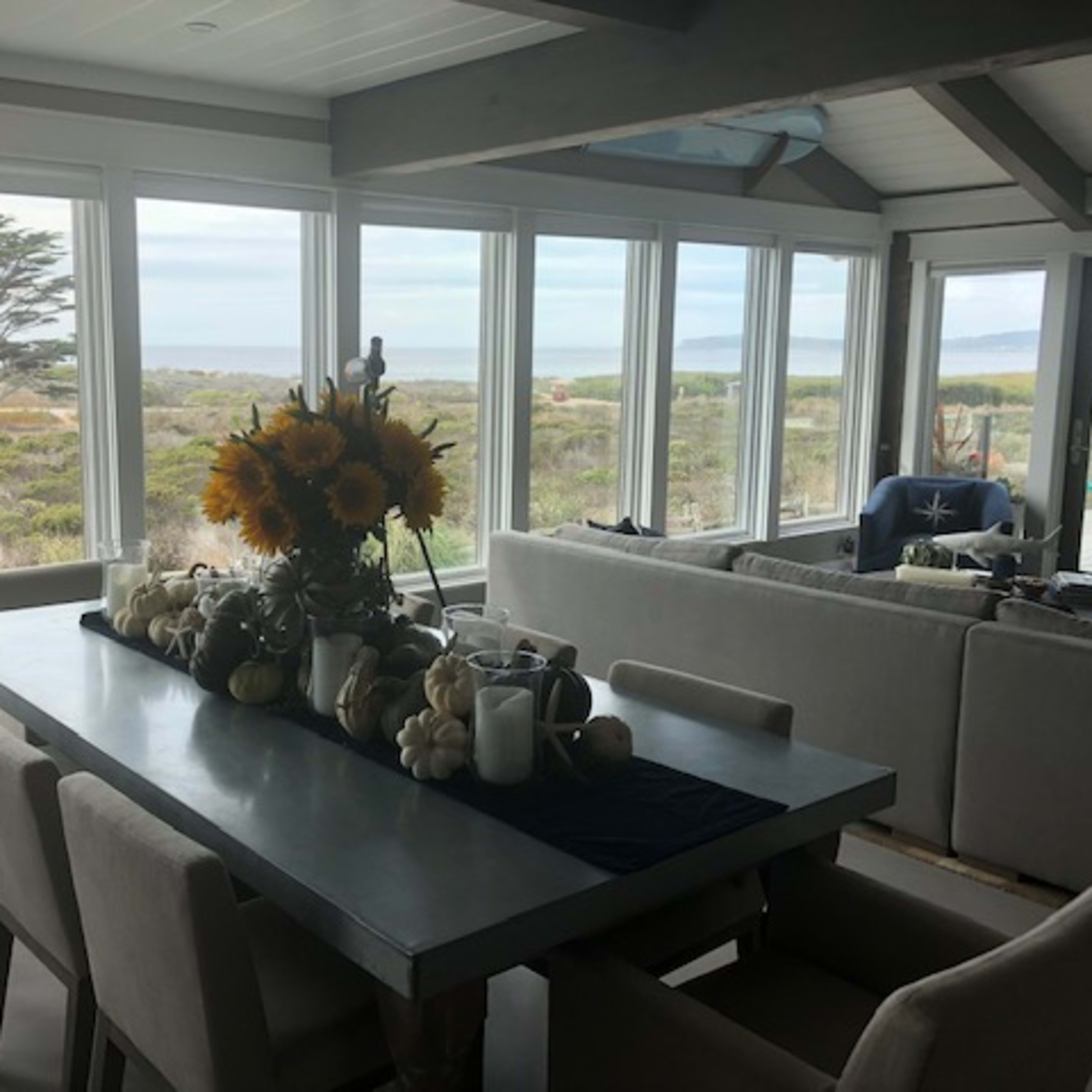 A dining area features a long table adorned with a flower arrangement and decorative items, surrounded by chairs and open windows that showcase a coastal view.