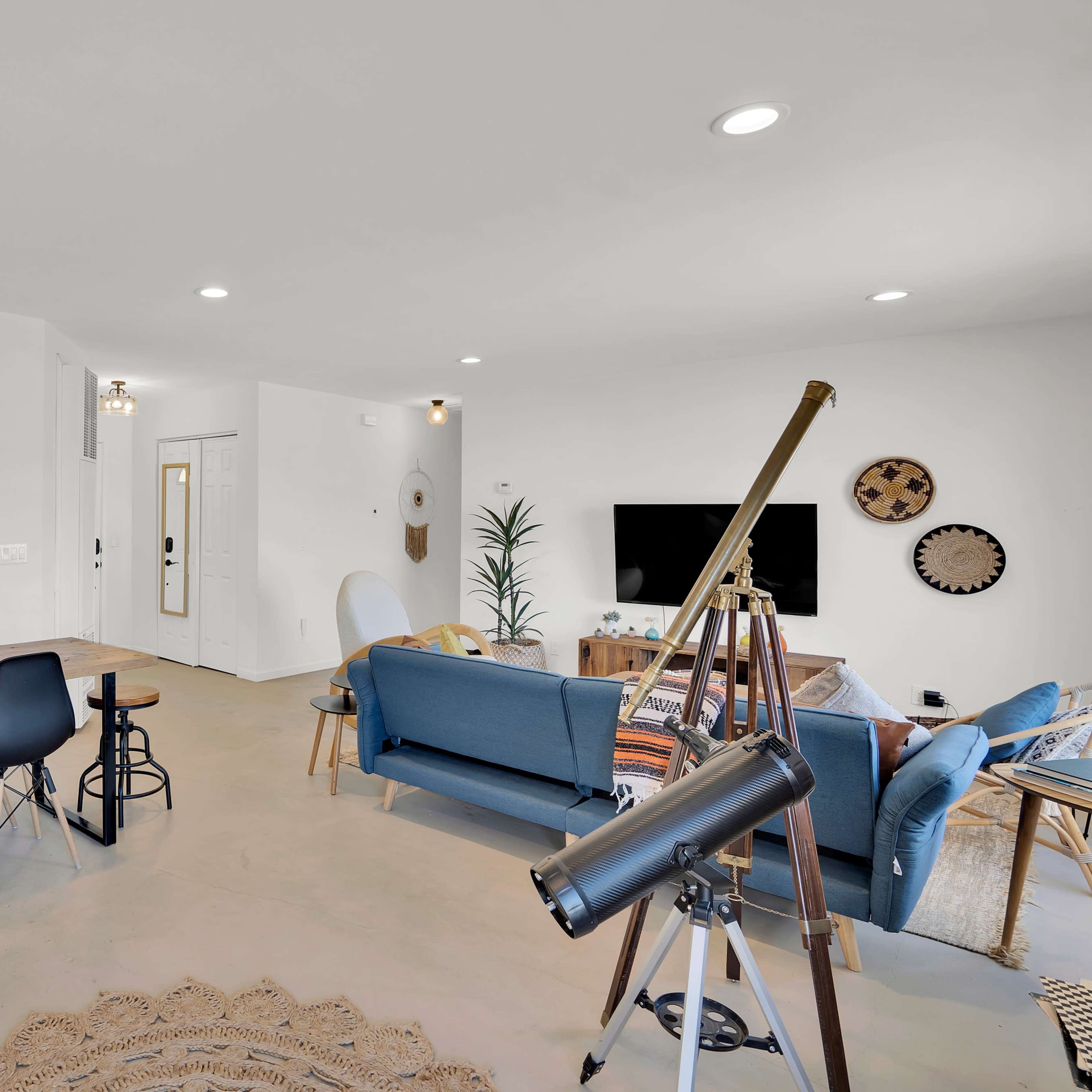 A modern, open-concept living space featuring a dining area with a wooden table and black chairs, a cozy seating area with blue couches, a large television, and a telescope.