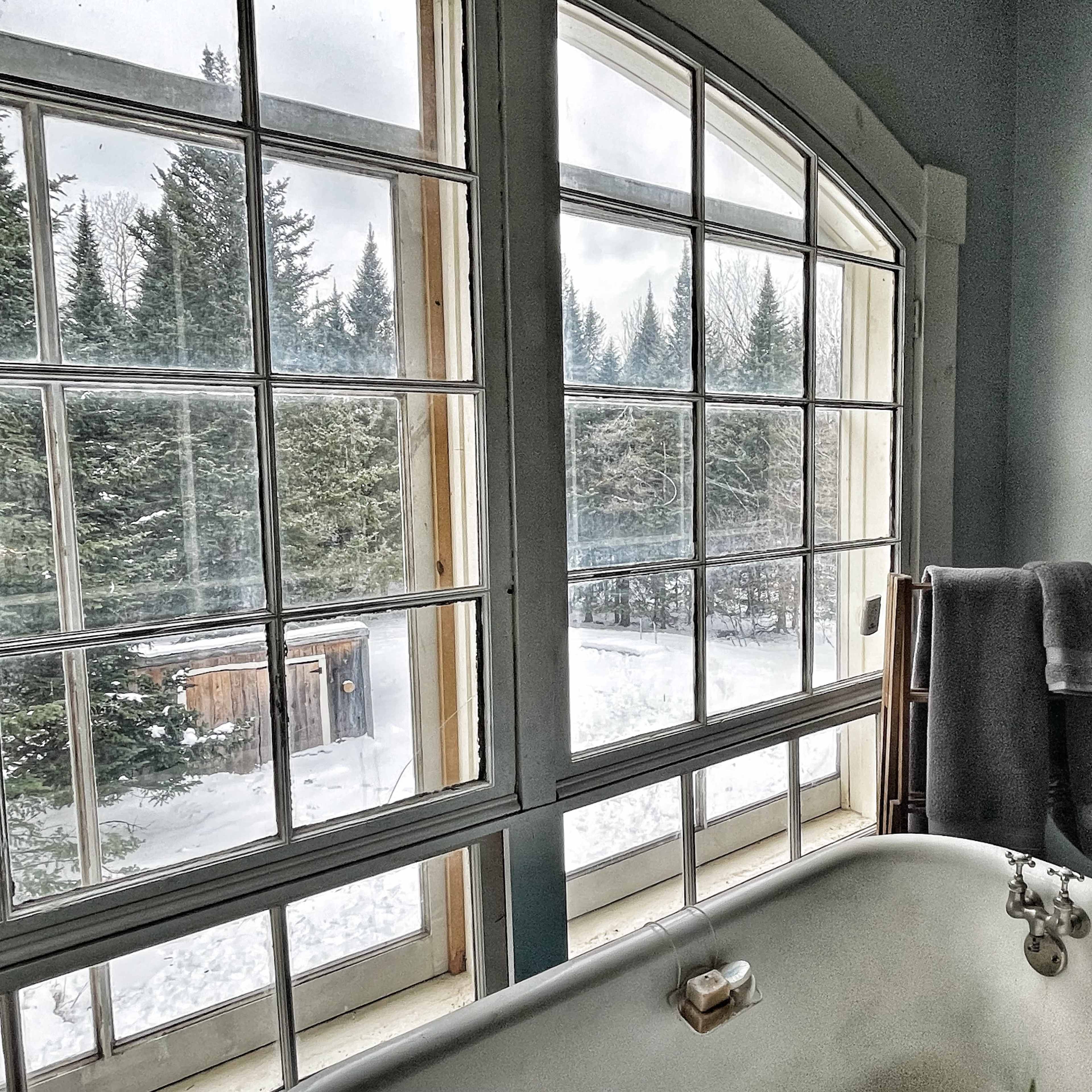 A vintage bathtub sits in a bathroom beside large windows that overlook a snowy landscape and evergreen trees.