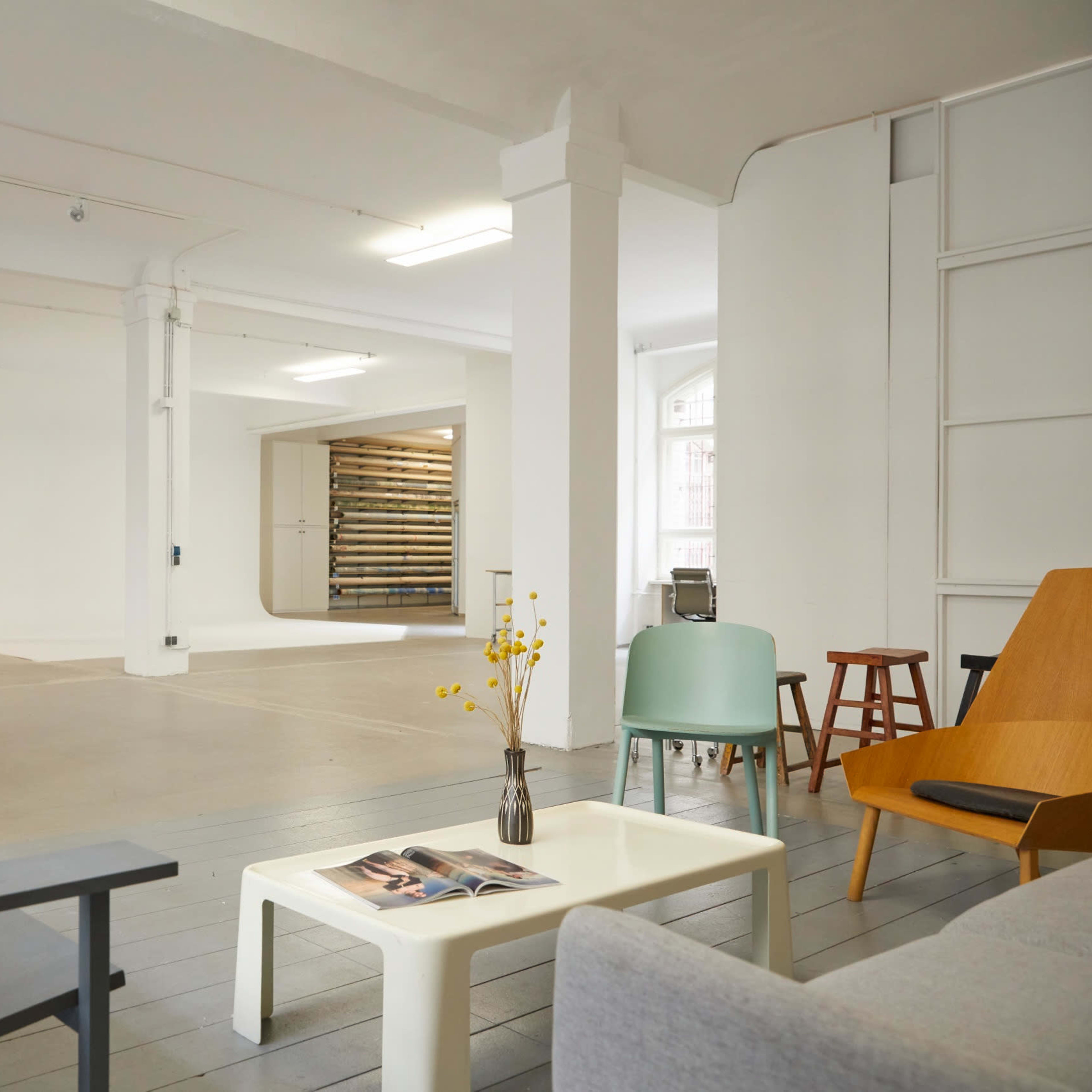A spacious, brightly lit room with a minimalist design featuring various seating arrangements, a dining table, and a wooden storage area in the background.