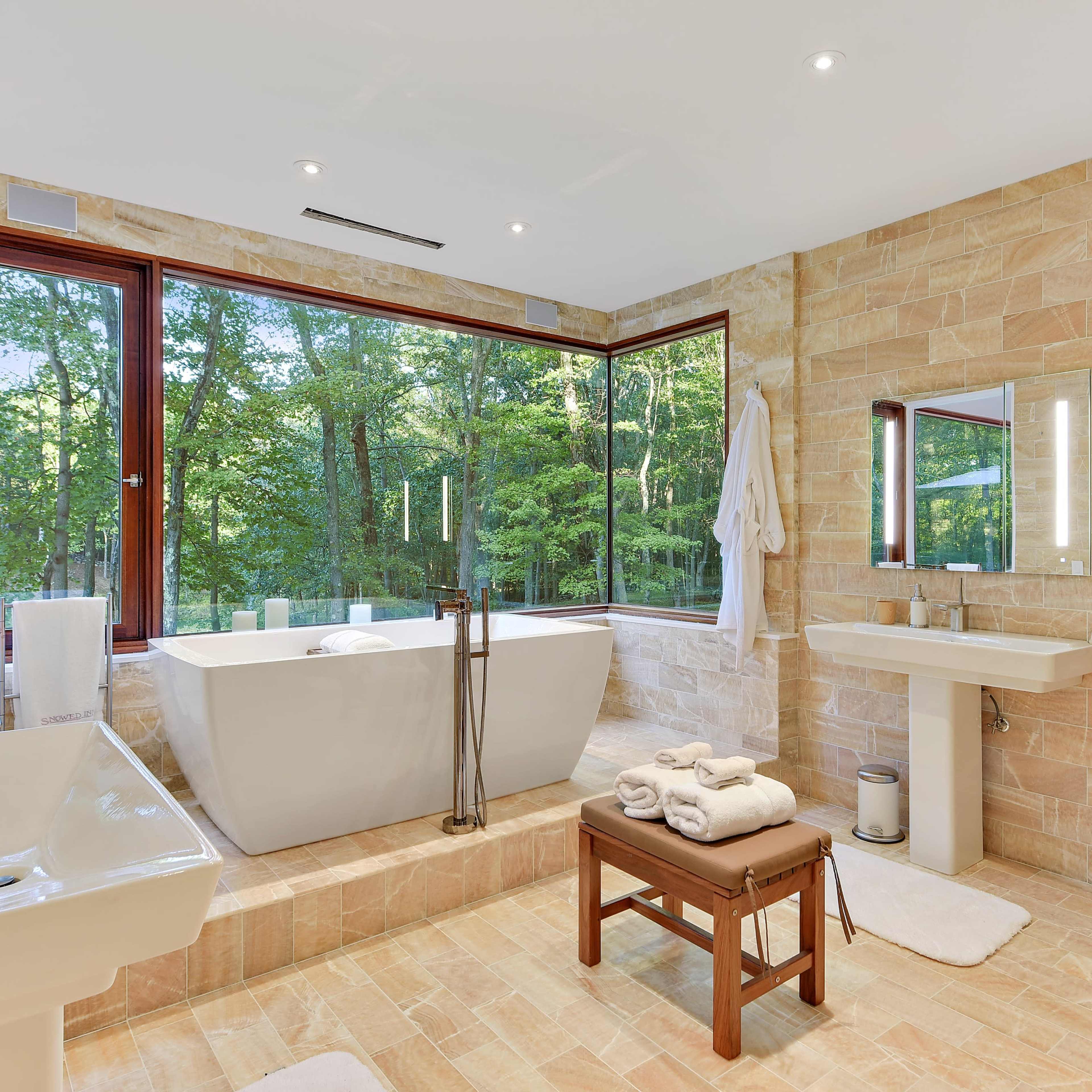 A spacious bathroom with a freestanding bathtub, a large window overlooking greenery, a sink, and a wooden bench with neatly rolled towels.
