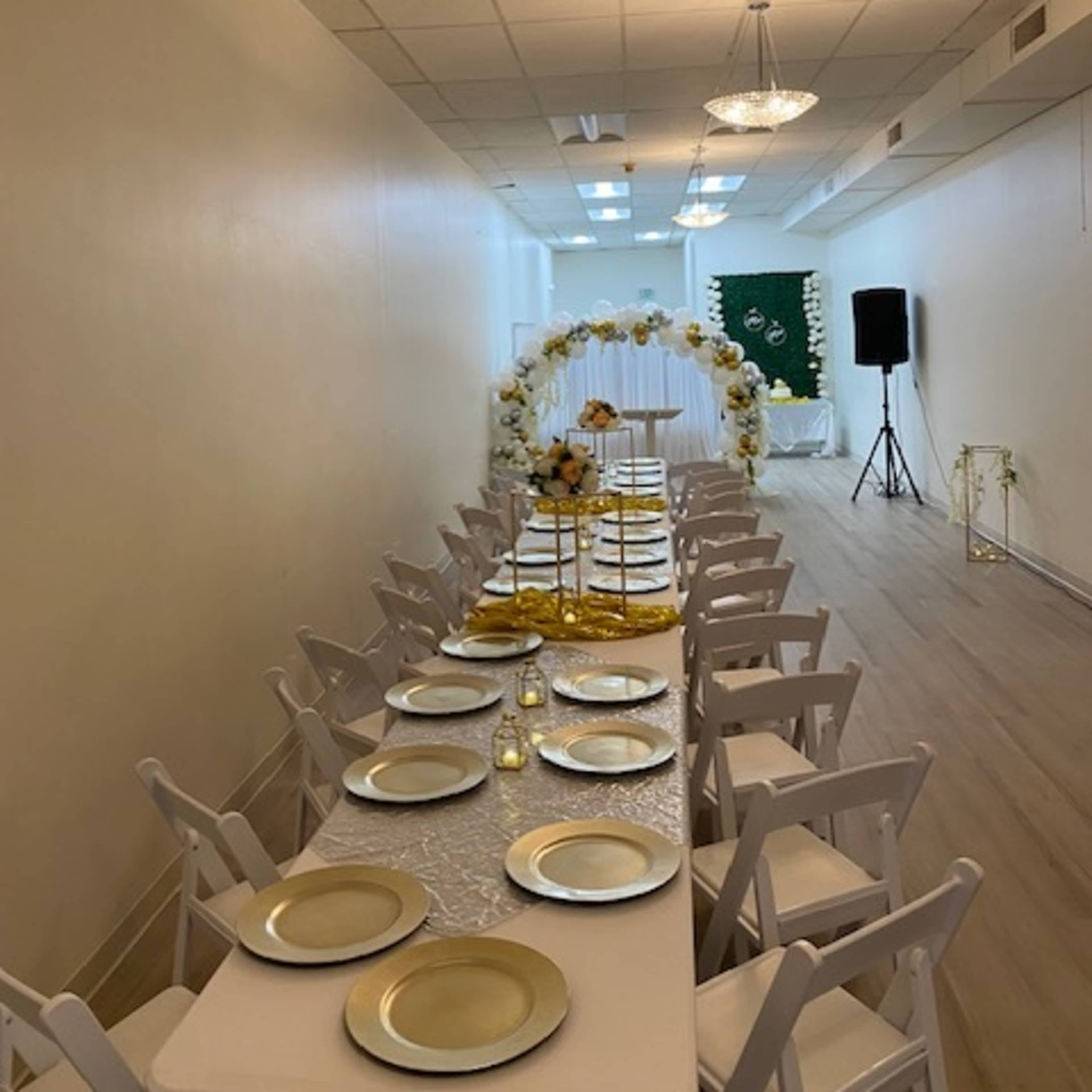 A long table set with golden plates is arranged in a narrow event space, leading to an arch decorated with flowers and a backdrop with green fabric.