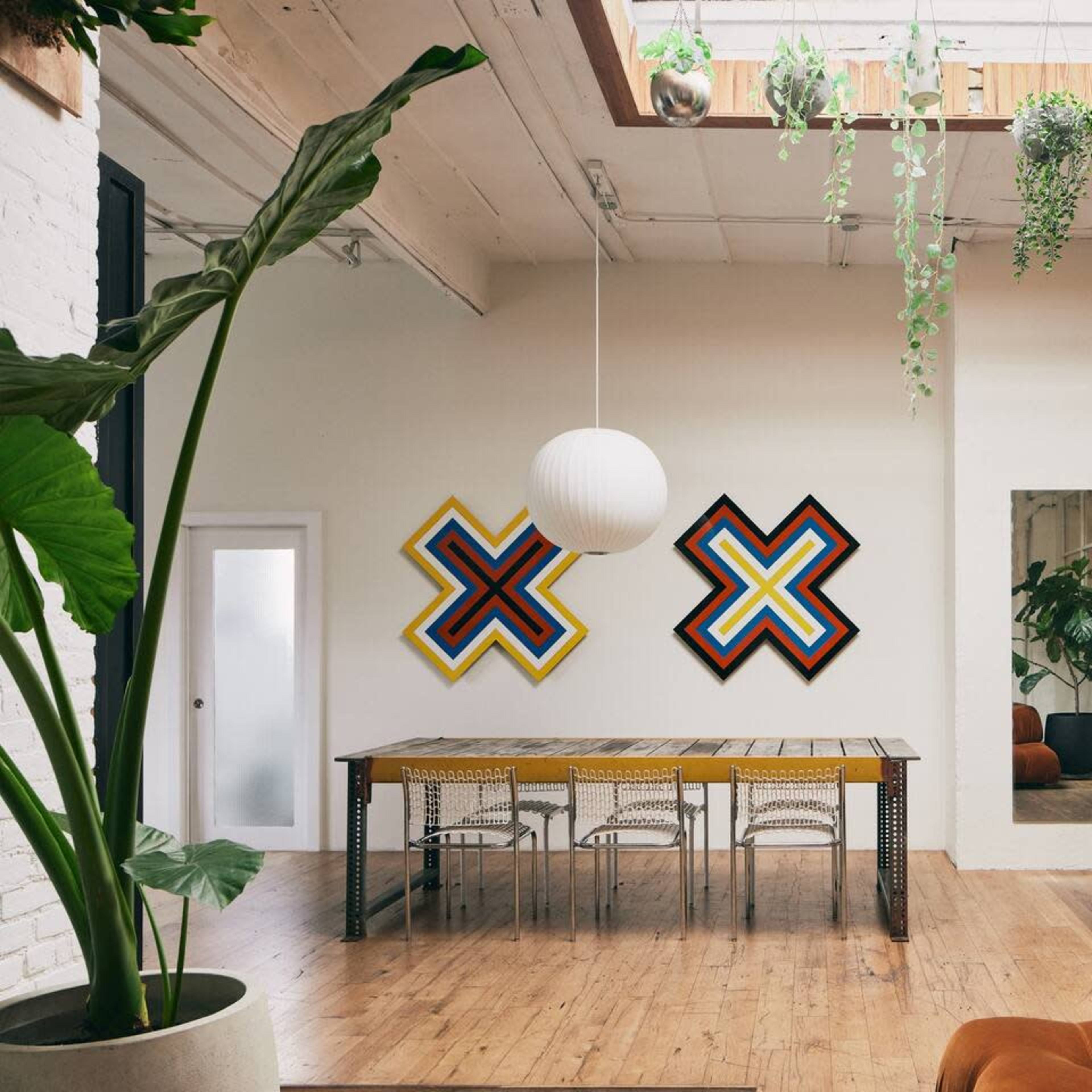 A spacious room with a wooden dining table surrounded by chairs, two colorful geometric artworks on the walls, a large mirror, and a motorcycle beside a whiteboard.