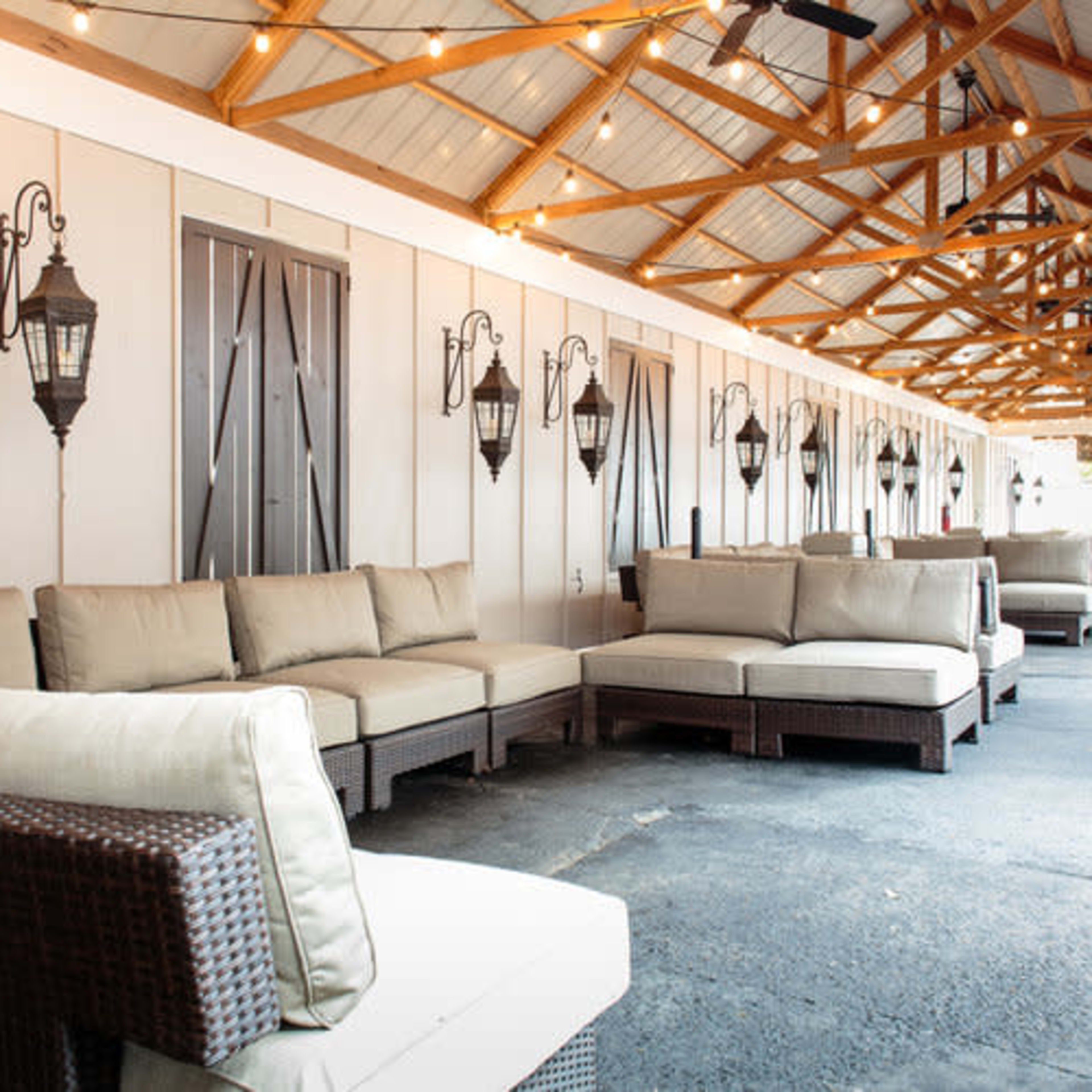 An outdoor lounge area with comfortable couches arranged under a wooden roof, illuminated by string lights and featuring wall-mounted lanterns.