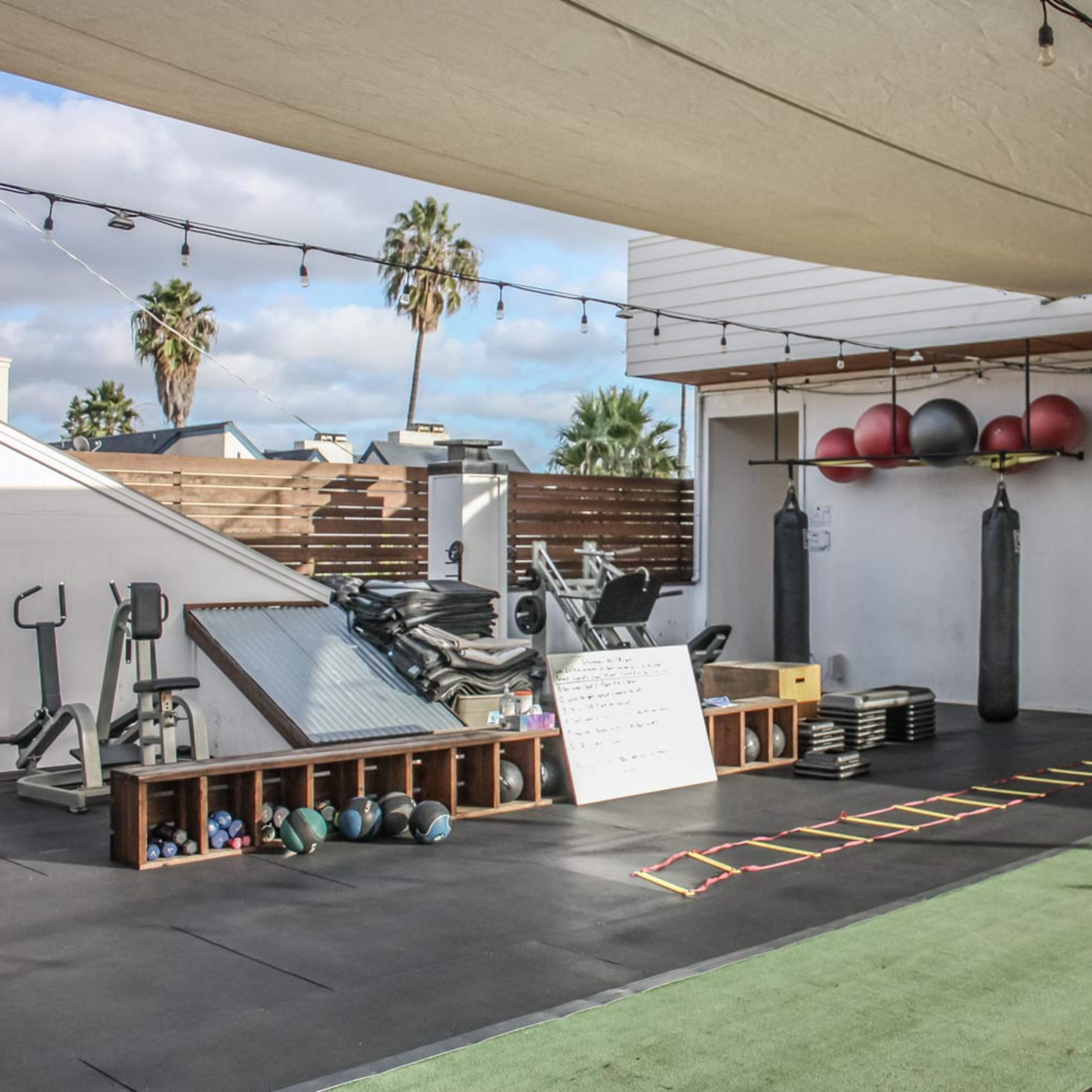 An outdoor gym setup with various exercise equipment, including weights, a mirror, and training tools, beneath a shaded area with palm trees in the background.