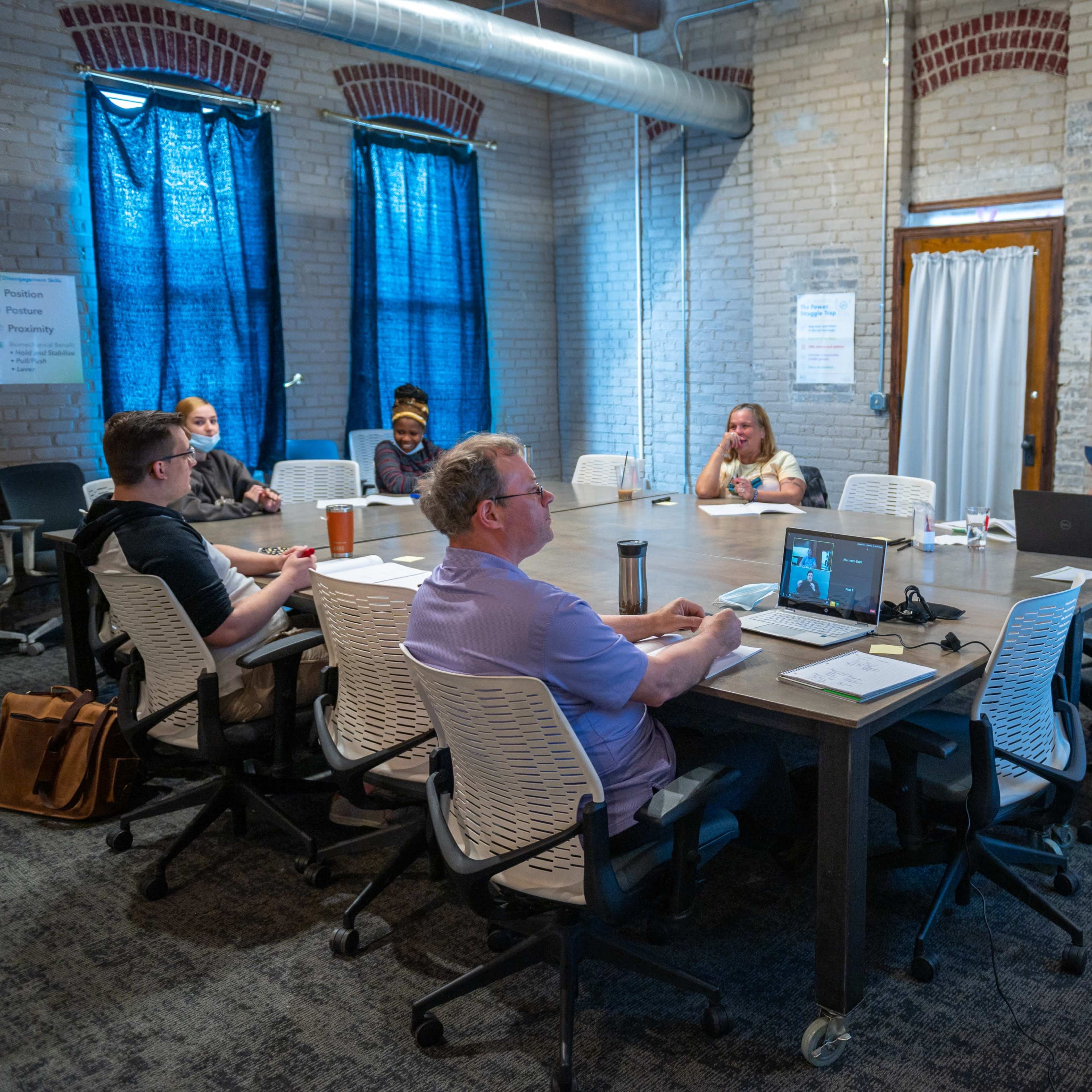 A group of six people sits around a large table in a conference room with exposed brick walls and large windows covered by blue curtains.
