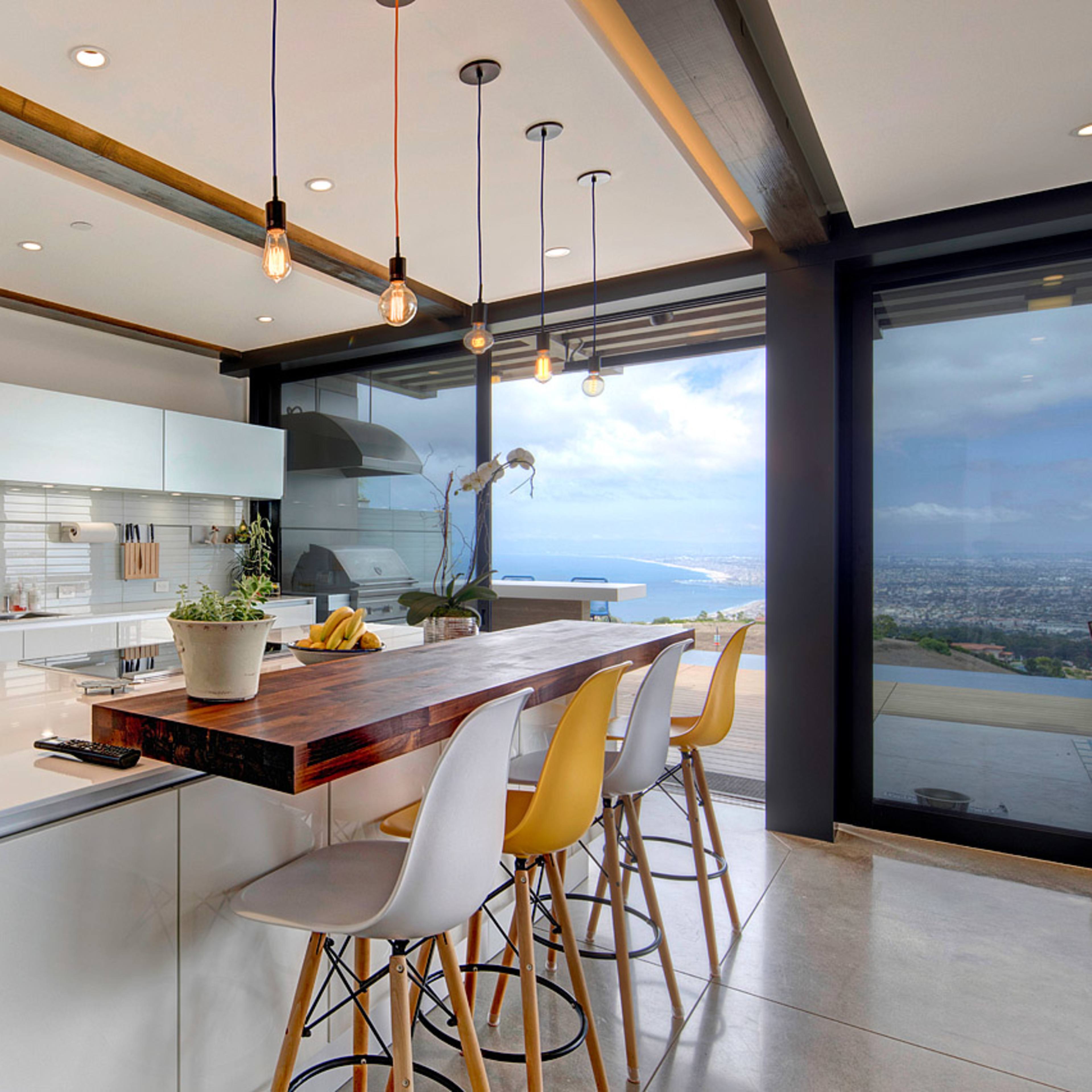 A modern kitchen with a large island, white cabinetry, and a panoramic view through glass walls.