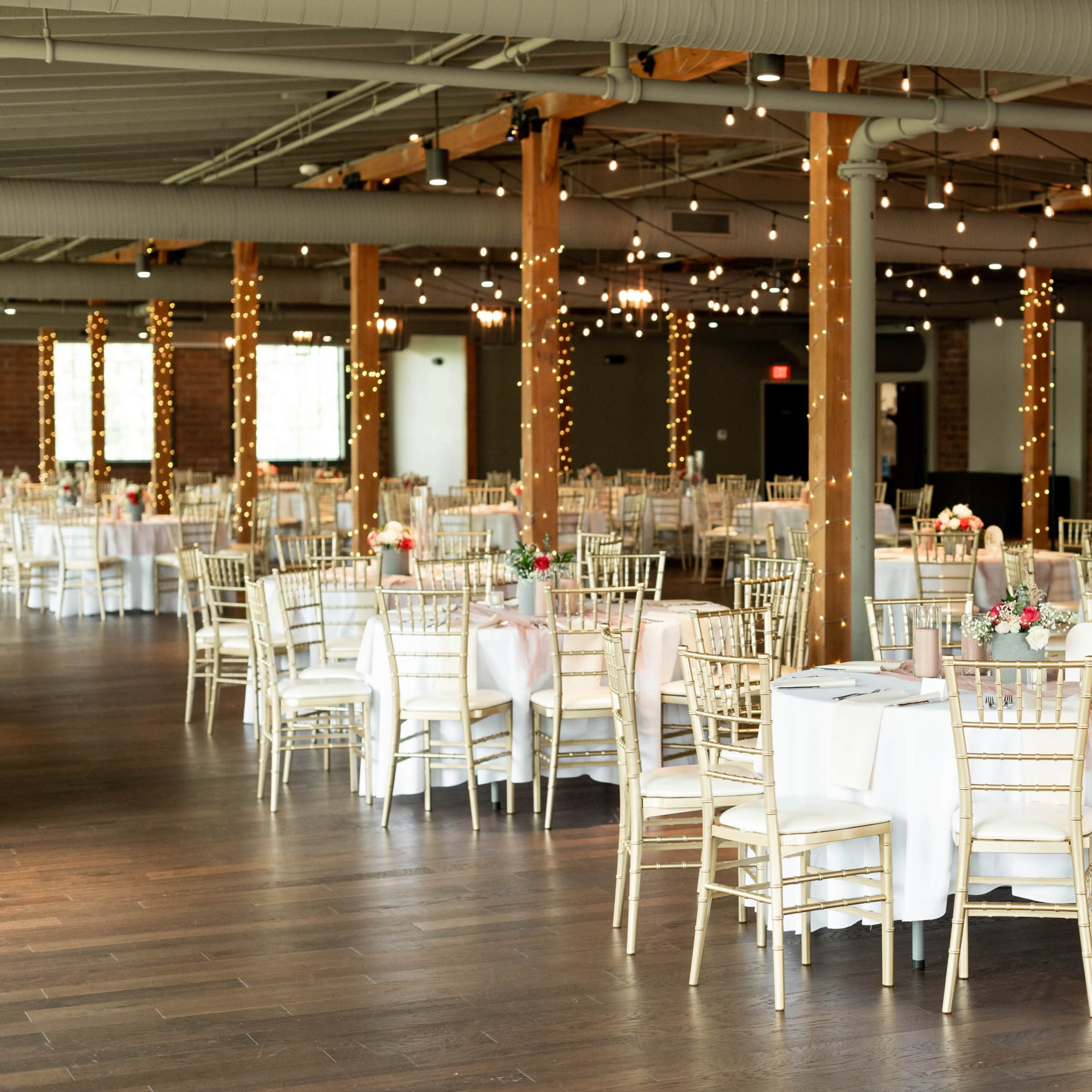 A spacious event venue features elegantly set tables with white tablecloths and gold chiavari chairs, illuminated by string lights and natural light from large windows.